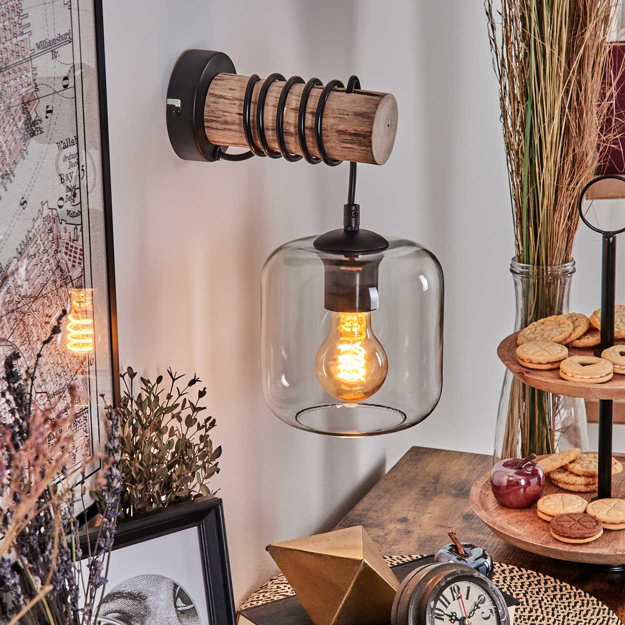 Eine Wandlampe mit Holzbügel und Glasschirm mit leuchtender Glühbirne. Kekse und Deko liegen auf einem Holztablett.