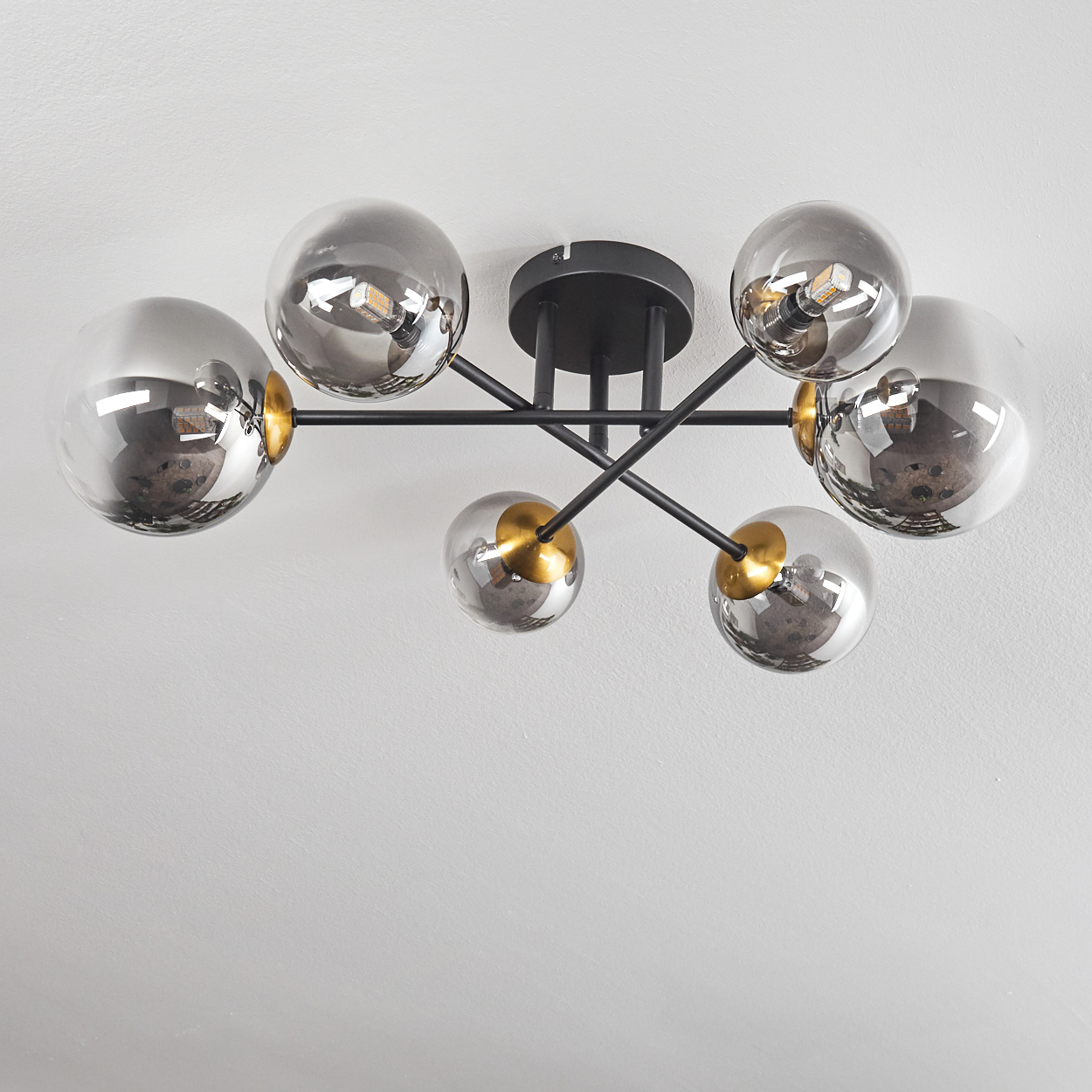 Eine moderne Deckenleuchte mit sechs grauen Glaskugeln. Die Leuchte hat ein schwarzes Metallgestell und goldfarbene Akzente. Sie ist an einer weißen Decke.