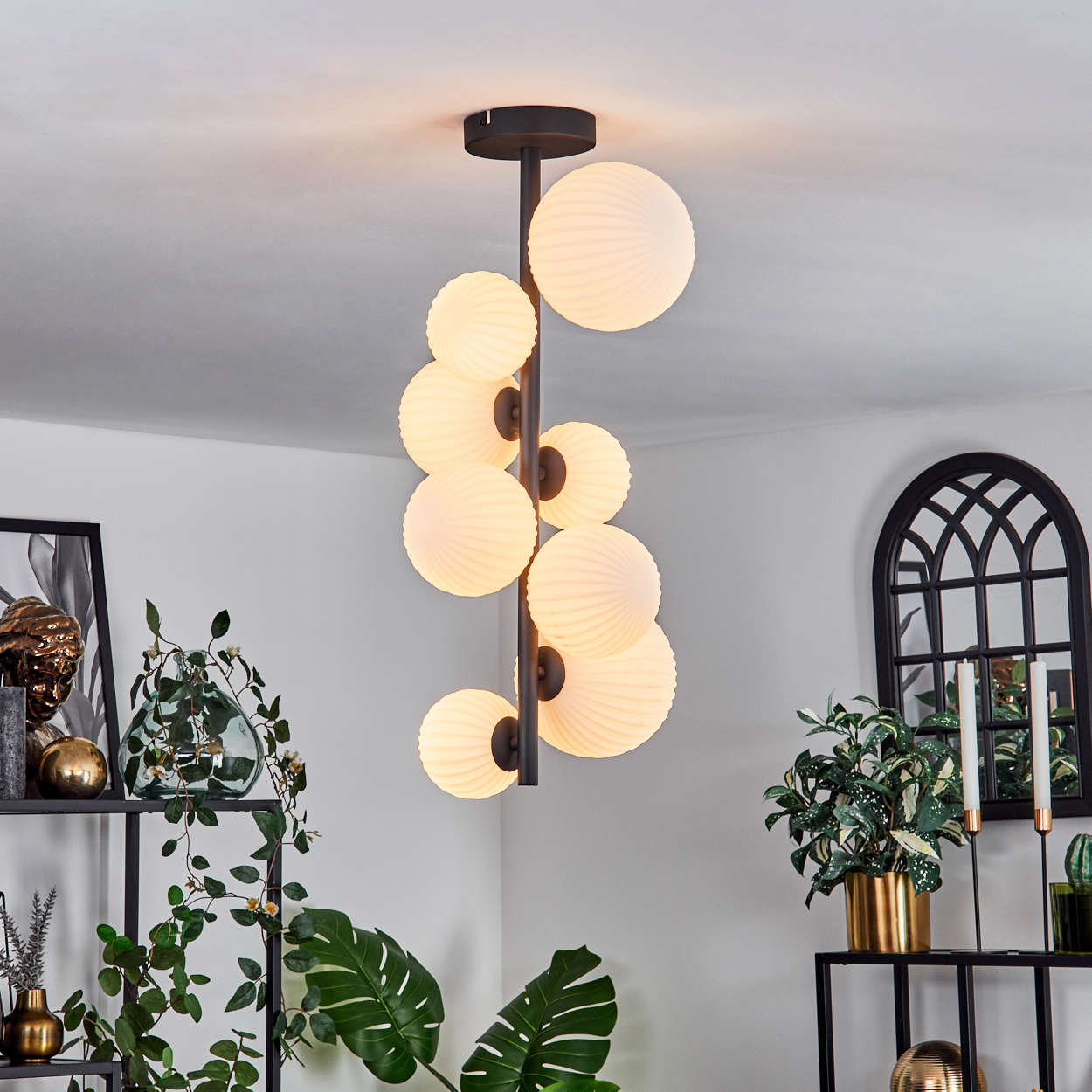 Eine moderne Hängelampe mit vielen kugelförmigen Lichtern hängt in einem Raum. Im Hintergrund befinden sich Pflanzen, Regale und ein bogenförmiger Spiegel.