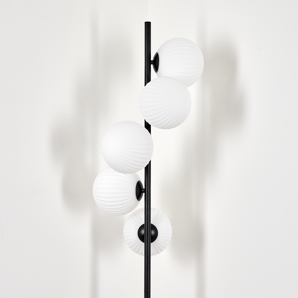 Eine schwarze Stehlampe mit fünf weißen, kugelförmigen Lampen. Die Lampen haben eine geriffelte Textur. Der Hintergrund ist eine weiße Wand.
