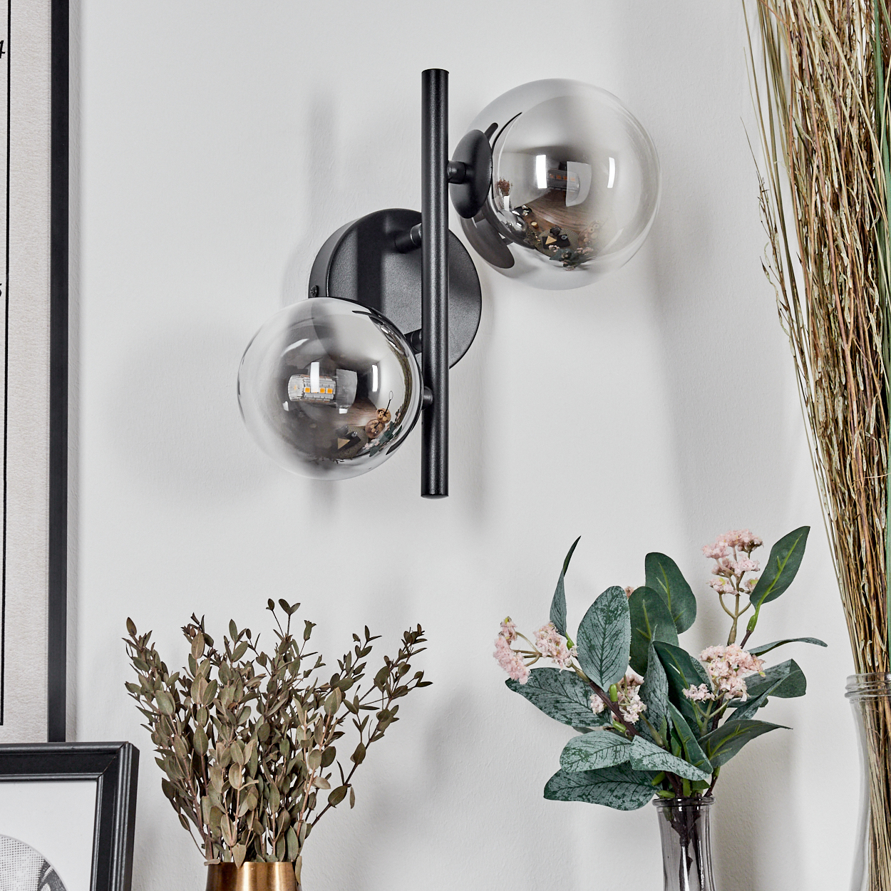Eine Wandlampe mit zwei Glaskugeln ist an einer weißen Wand befestigt. Getrocknete Pflanzen stehen in Vasen darunter, ein gerahmtes Bild links.