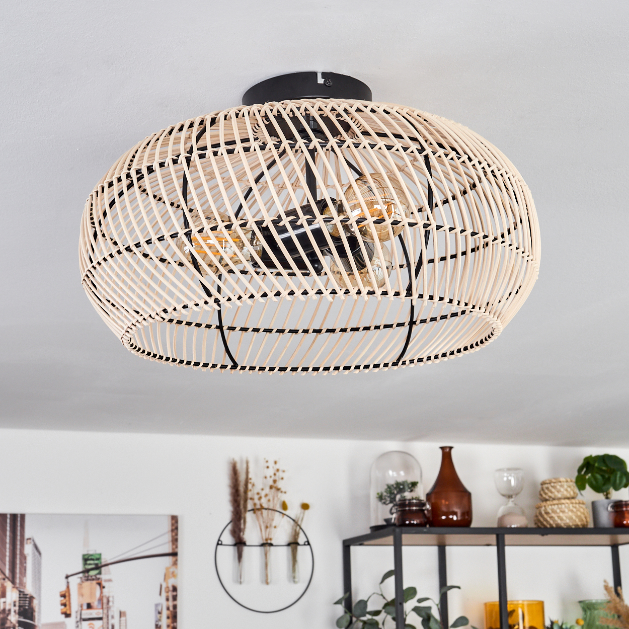 Eine Deckenlampe aus gewebtem Rattan mit schwarzen Details. Im Hintergrund Regale mit Deko und eine Wand mit Bild und Pflanzen.