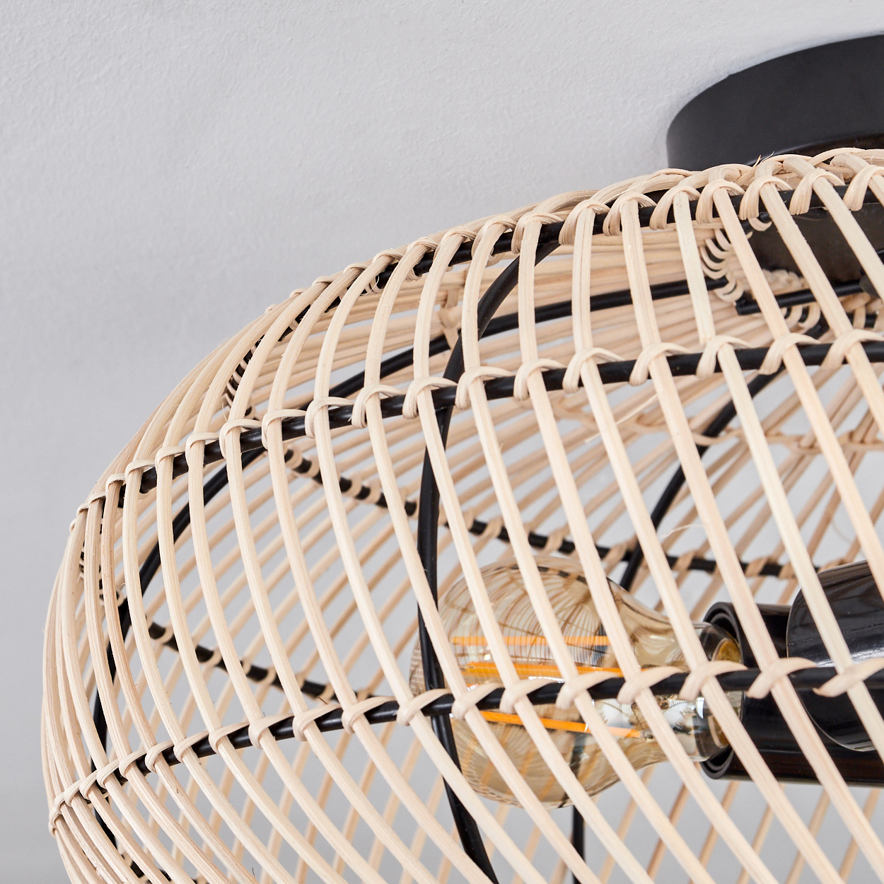 Nahaufnahme einer gewebten Rattanlampe mit schwarzem Metallrahmen. Im Inneren befindet sich eine Glühbirne. Der Hintergrund ist eine weiße Decke.
