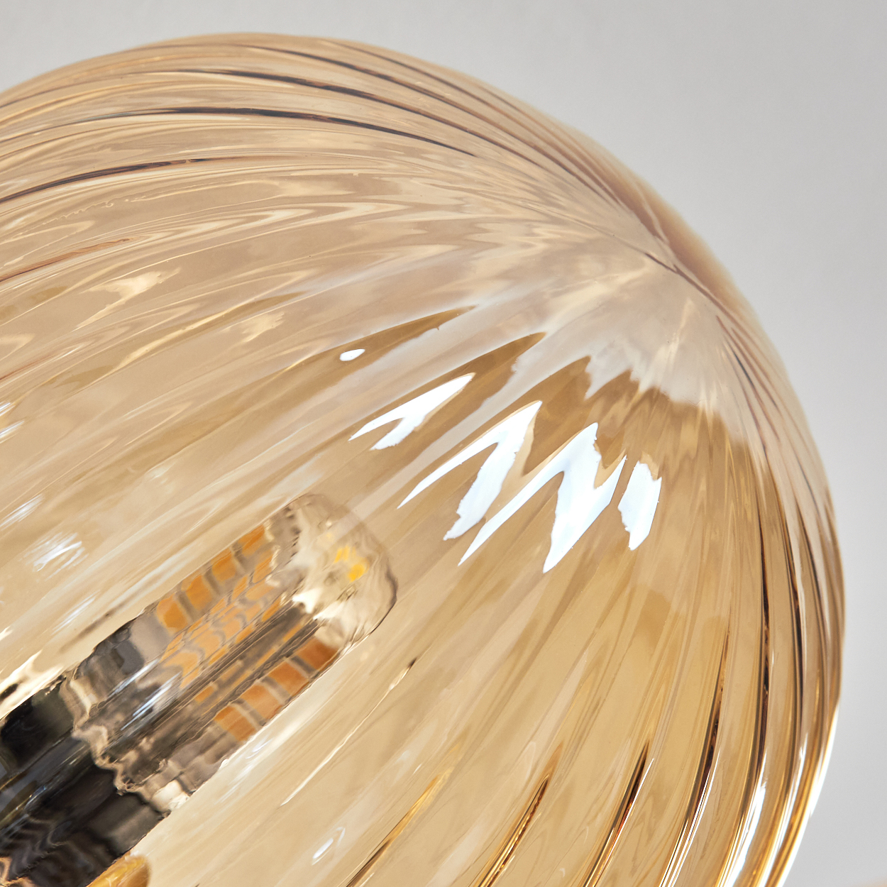 Nahaufnahme einer Leuchte. Sie hat einen goldenen, gerippten Glasschirm. Die Glühbirne ist im Inneren des Schirms sichtbar. Der Hintergrund ist weiß.