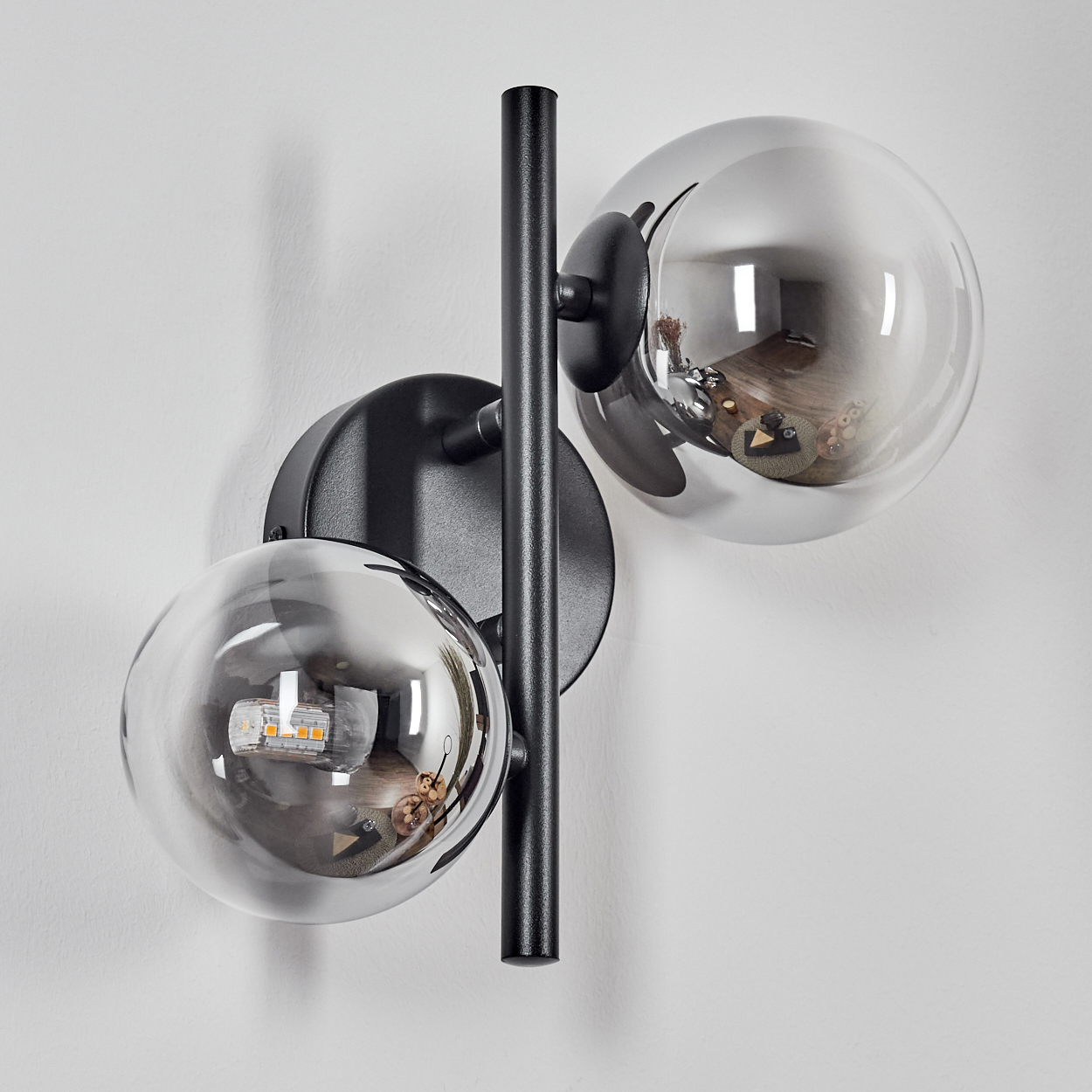 Schwarze Wandlampe mit zwei Glaskugeln. Die Kugeln reflektieren einen Raum mit Objekten. An einer weißen Wand montiert.