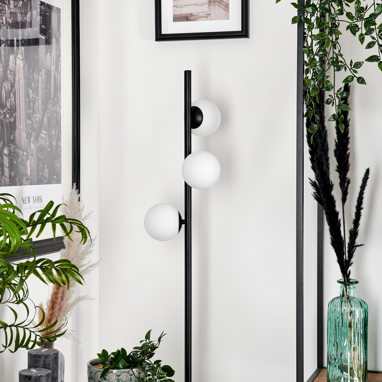 Eine schwarze Stehlampe mit drei weißen Kugelleuchten. Pflanzen und gerahmte Bilder schmücken die weiße Wand im Hintergrund.