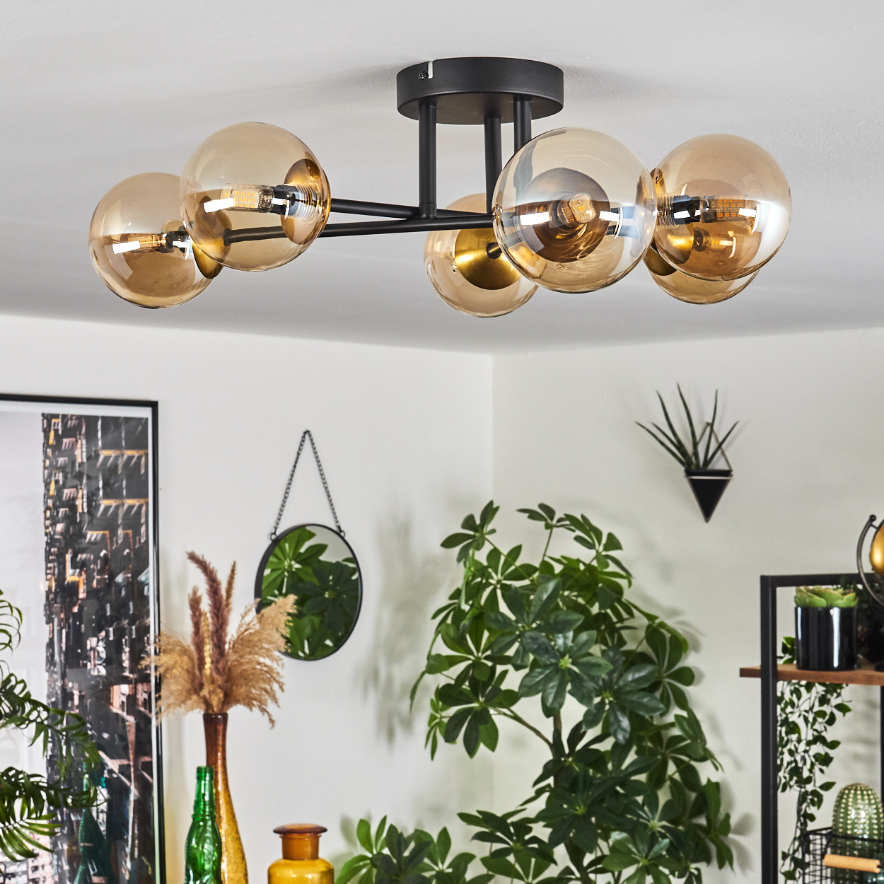 Eine moderne Deckenleuchte mit sechs bernsteinfarbenen Glaskugeln ist an einer weißen Decke befestigt. Pflanzen und Dekor sind im Hintergrund.