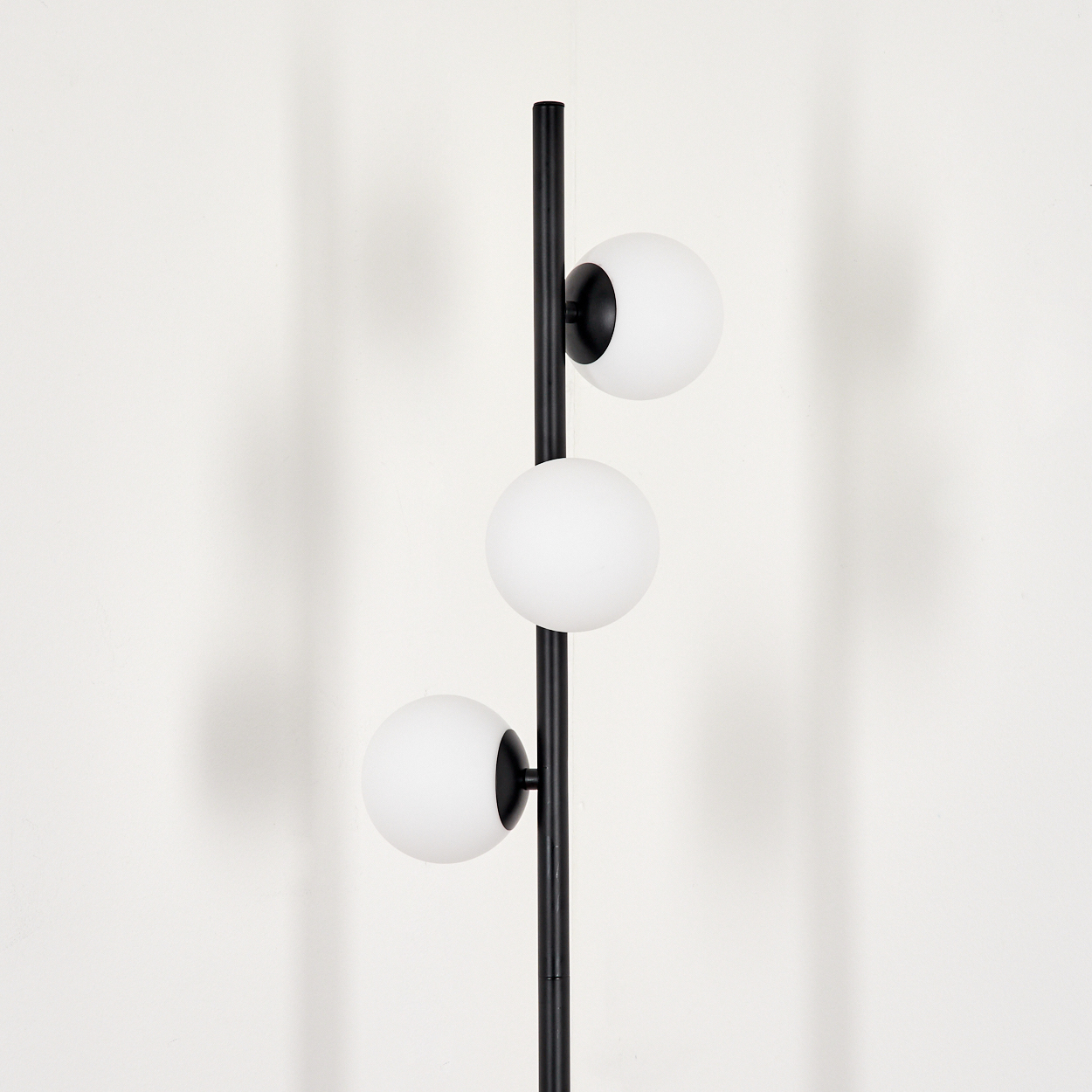 Eine schwarze Stehlampe mit drei weißen Kugelleuchten. Die Lampe steht vor einer weißen Wand.