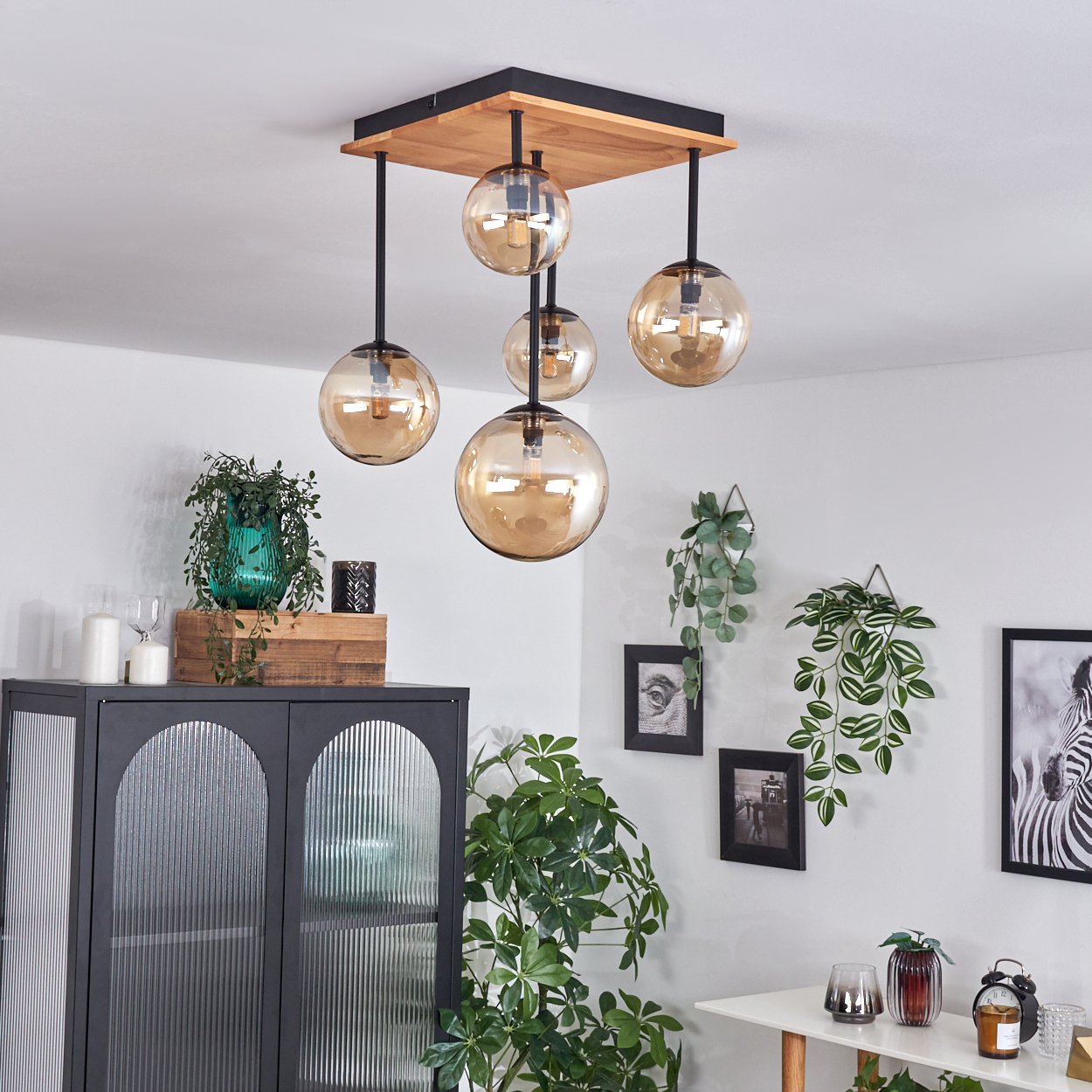 Moderne Deckenlampe mit fünf kugelförmigen Glasschirmen. Pflanzen, ein Schrank und gerahmte Bilder schmücken die weißen Wände.