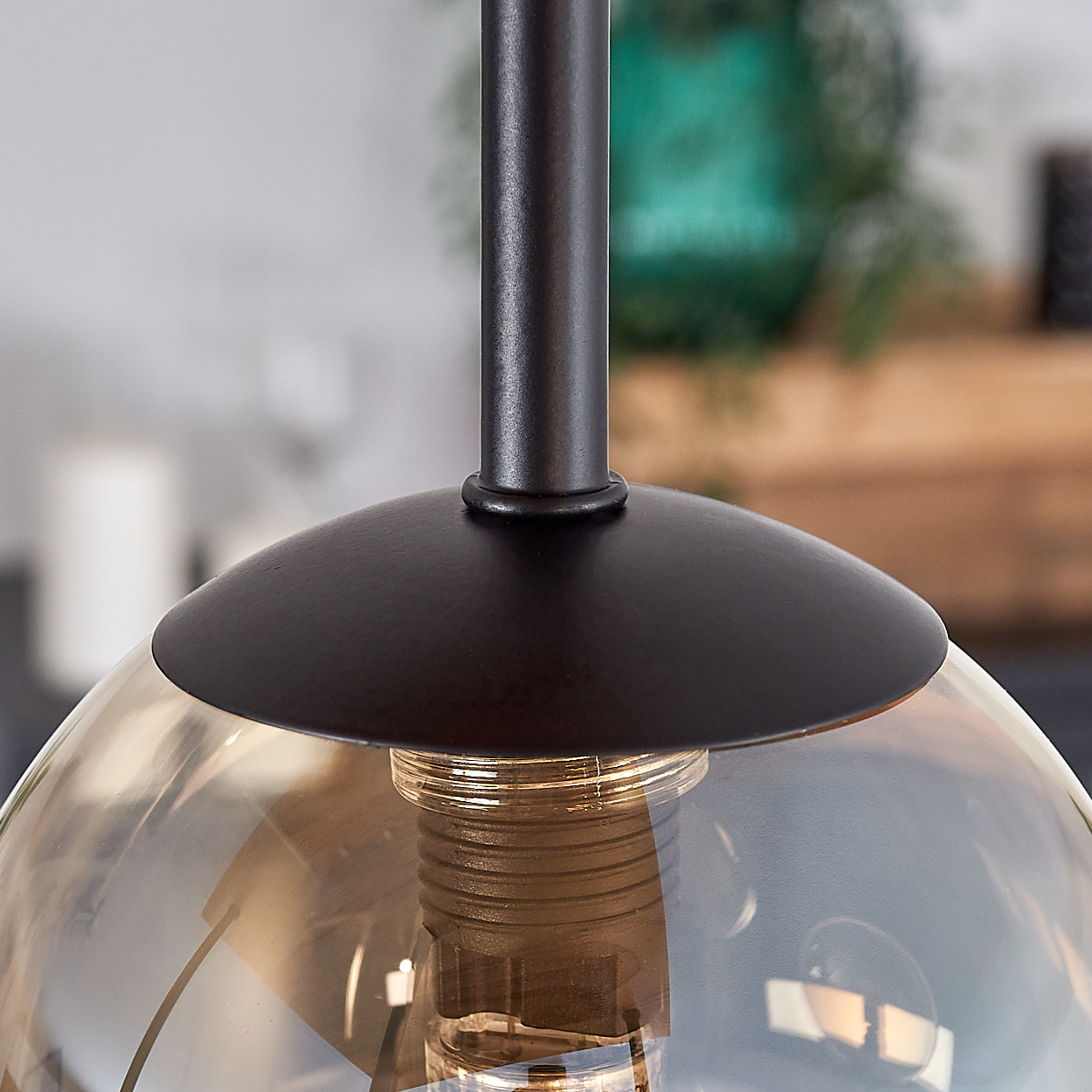 Nahaufnahme einer Pendelleuchte. Eine dunkle Metallhalterung trägt einen Glasschirm. Die Glühbirne ist im Glas sichtbar. Der Hintergrund ist unscharf.