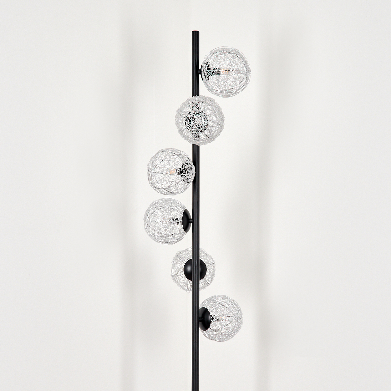 Eine schwarze Stehlampe mit sechs kugelförmigen Drahtrahmen-Leuchten steht vor einer weißen Wand. Die Leuchten sind silberfarben.