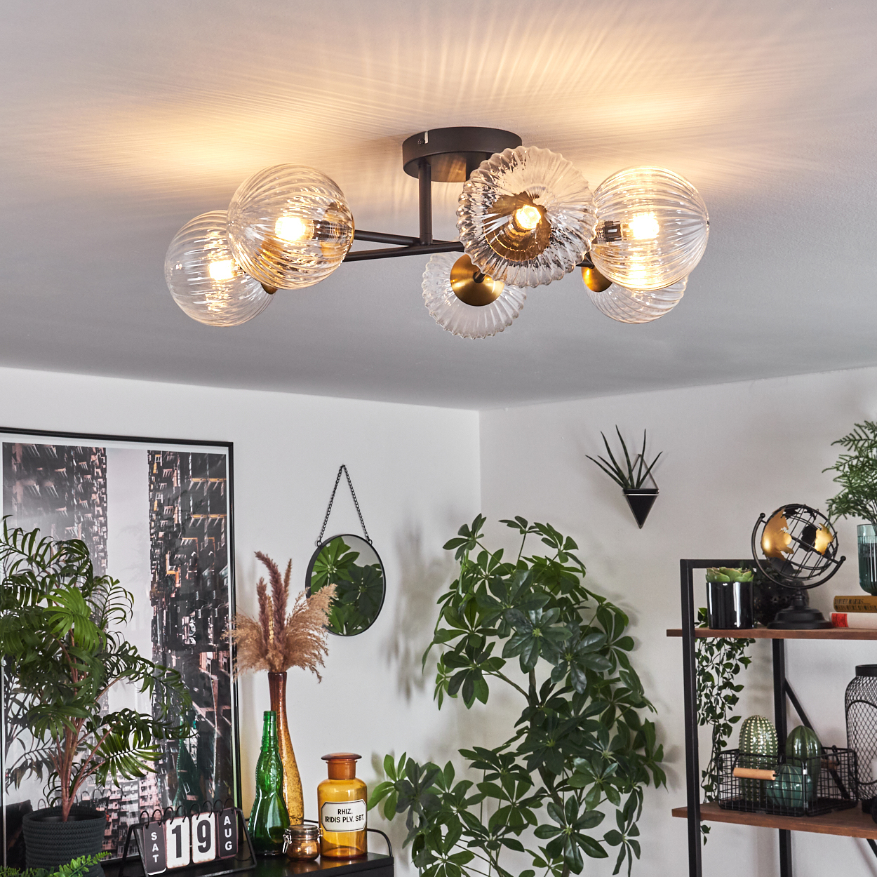 Eine Deckenleuchte mit Glaskugeln. Darunter Pflanzen, Dekorationen und ein Regal mit Gegenständen. Ein Spiegel hängt an der Wand. Die Szene ist gut beleuchtet.