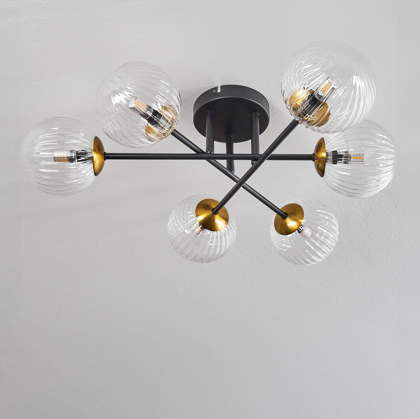 Moderne Deckenleuchte mit sechs Glaskugeln, schwarzen Metallarmen und goldenen Akzenten, an einer weißen Decke montiert.