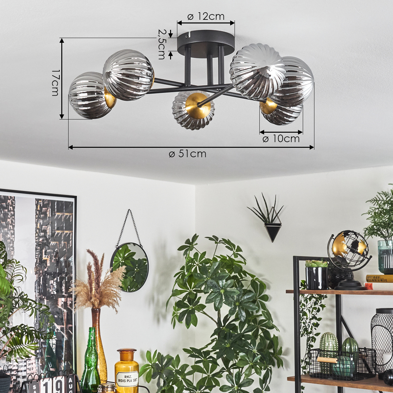 Eine Deckenlampe mit fünf silbernen Kugeln, schwarzen Armen und einem goldenen Zentrum. Im Hintergrund befinden sich Pflanzen, ein Spiegel und Regale mit Dekoration.