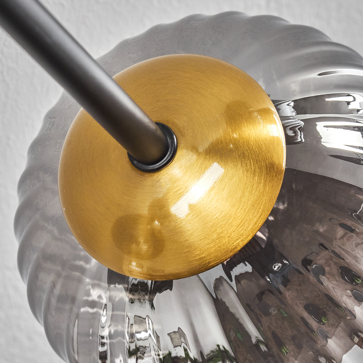 Nahaufnahme einer goldenen, runden Leuchte mit schwarzem Arm. Ein grauer, gewellter Glasschirm umgibt die Leuchte.