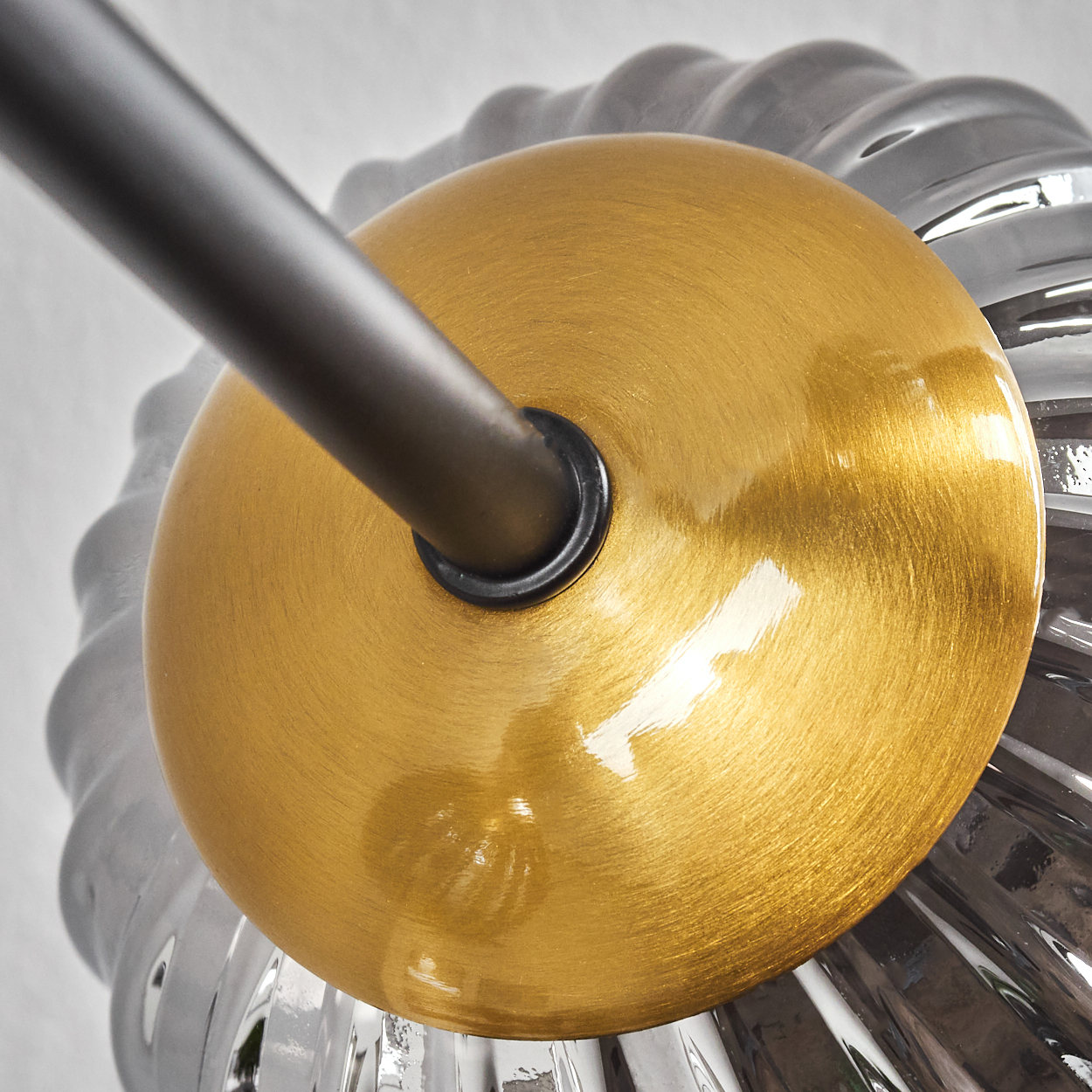 Nahaufnahme einer goldenen, kreisförmigen Lampenhalterung mit einer dunklen Stange, die sich davon erstreckt. Ein gerippter, transparenter Glasschirm ist sichtbar.