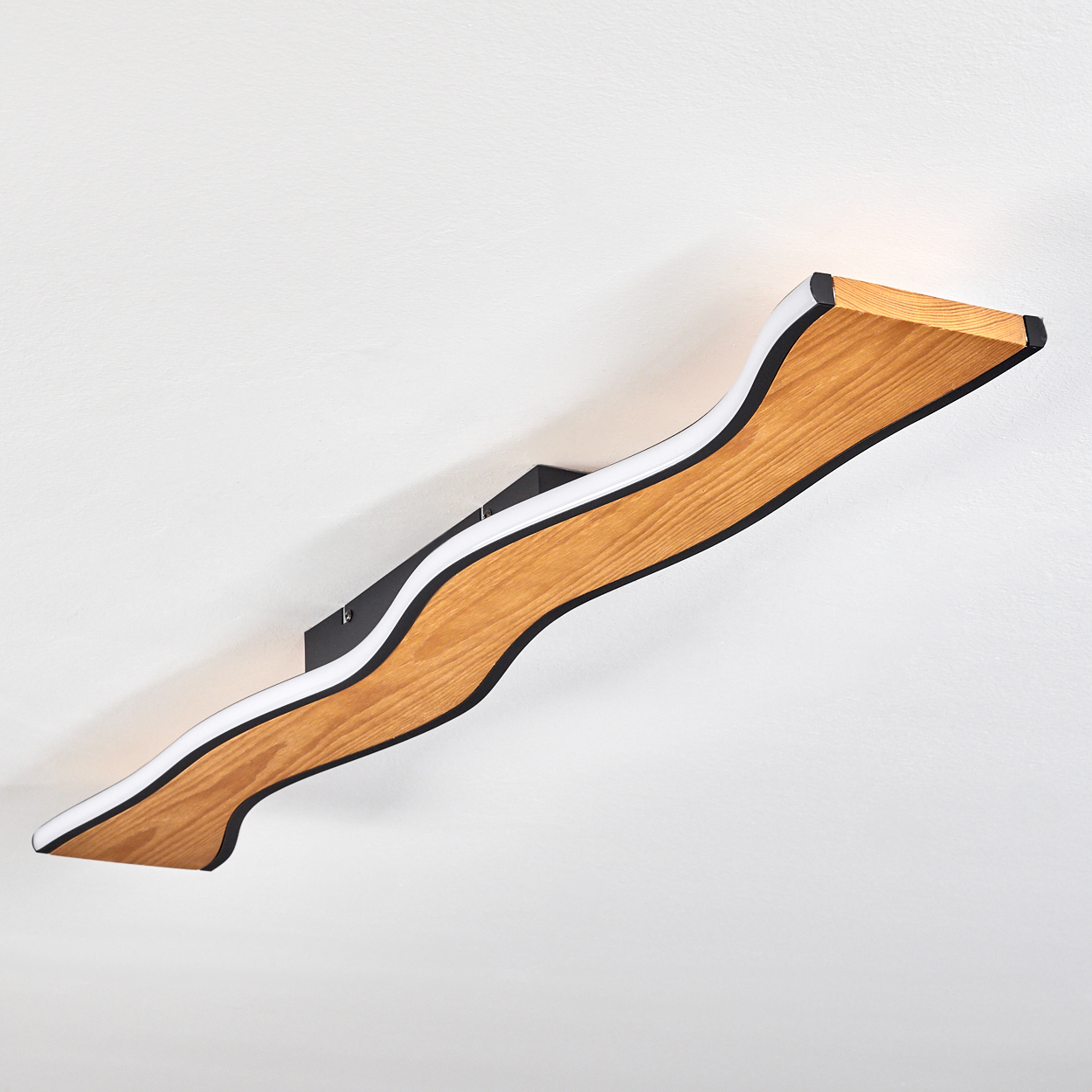 Eine holzfarbene, wellenförmige Wandlampe mit schwarzen Kanten und einem weißen Innenlicht. Sie ist an einer weißen Wand befestigt.