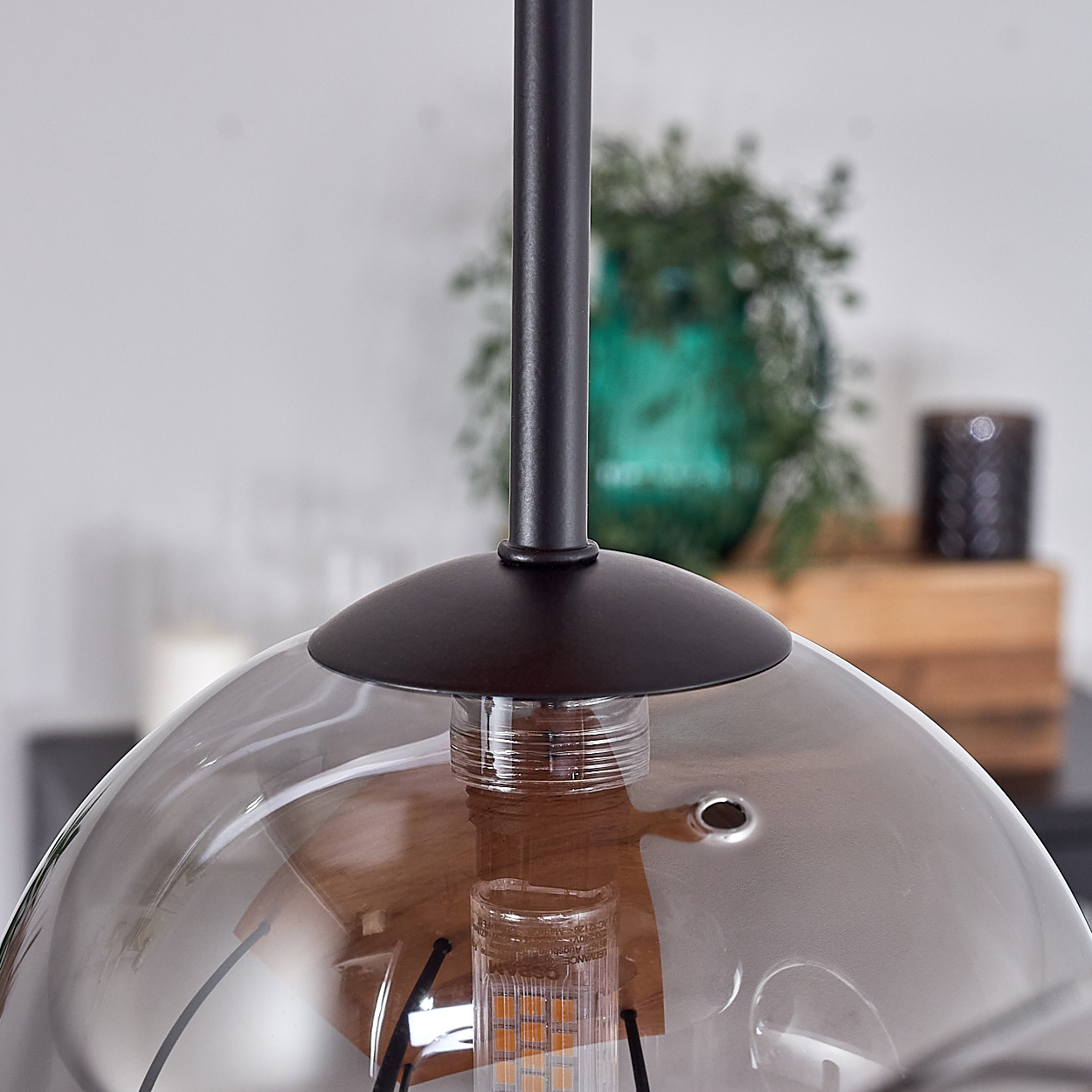 Eine moderne Pendelleuchte mit rauchfarbenem Glasschirm. Im Hintergrund sind eine grüne Vase und ein dunkler Kerzenhalter zu sehen.