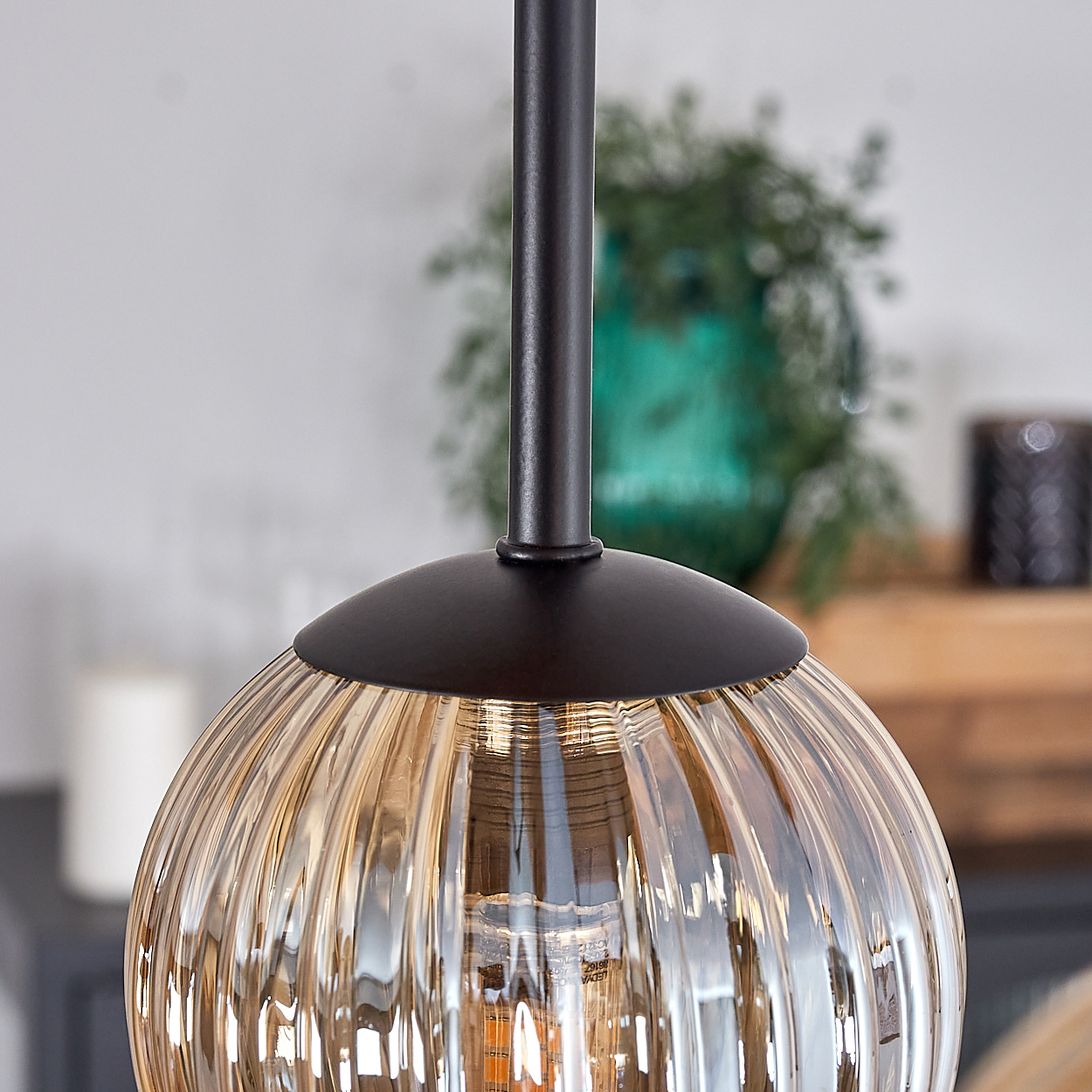 Nahaufnahme einer gerippten Glas-Hängeleuchte. Die Lampe ist braun und hat einen schwarzen Stiel. Der Hintergrund ist verschwommen mit grünen Pflanzen.