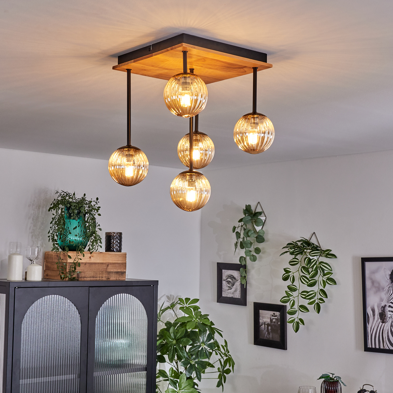 Eine moderne Deckenlampe mit fünf Kugelleuchten hängt an einer Holzdecke. Pflanzen, Bilder und ein Schrank befinden sich im Raum.