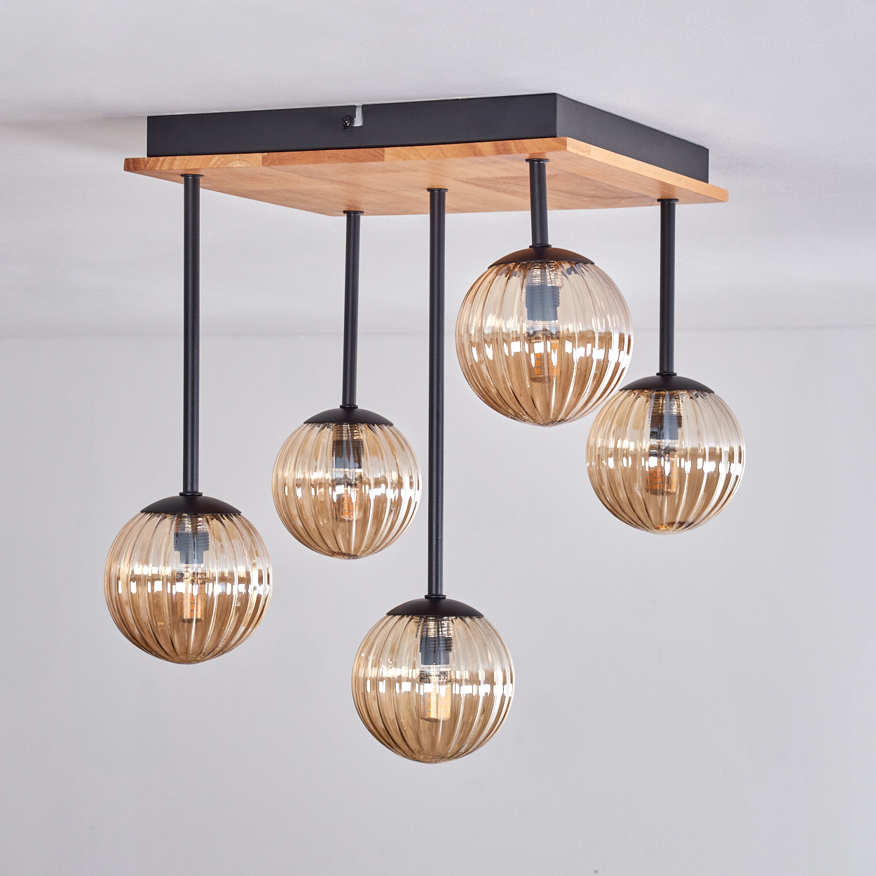Eine Deckenleuchte mit fünf bernsteinfarbenen Glaskugeln, die von einem Holz- und schwarzen Metallrahmen hängen. Der Hintergrund ist eine weiße Decke.