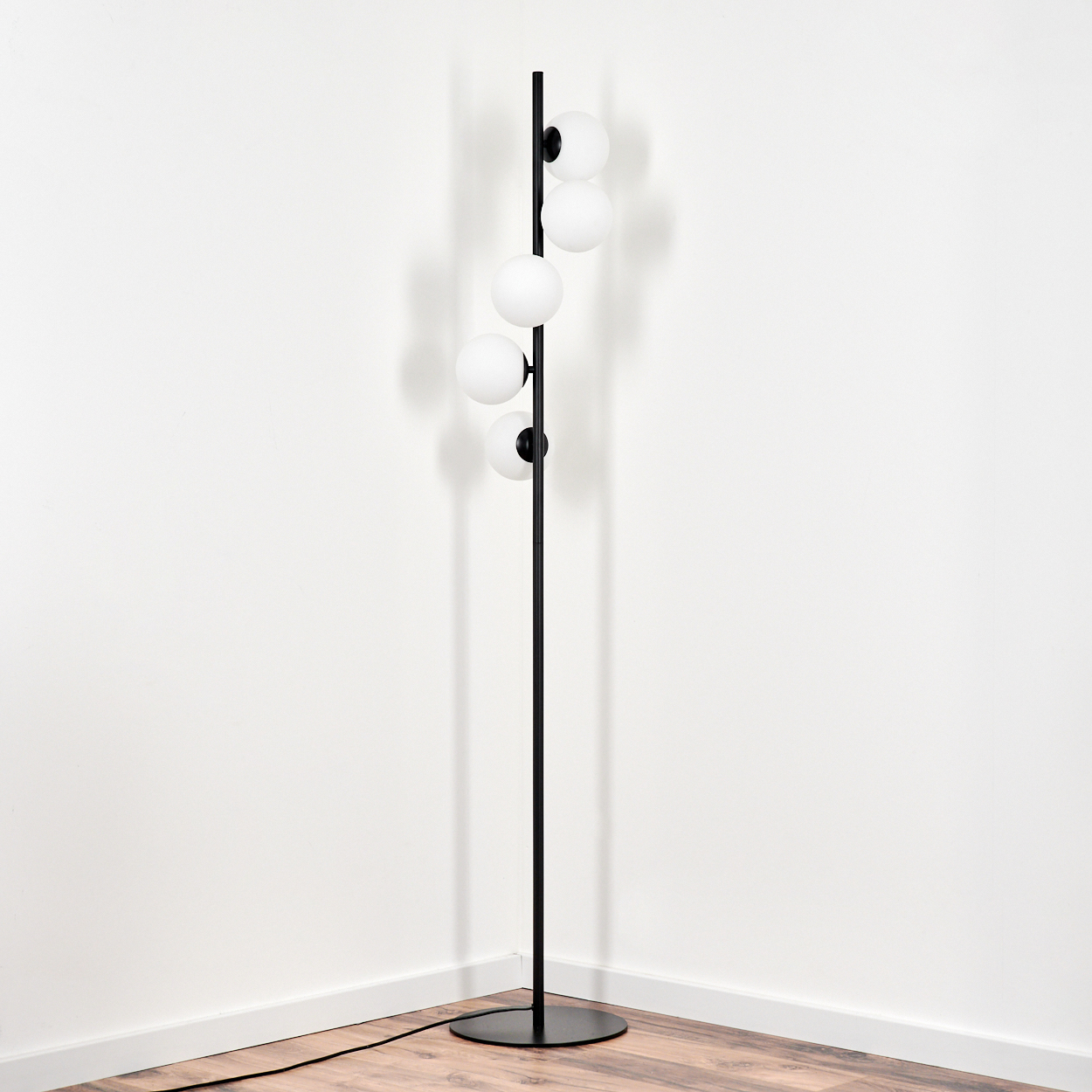 Eine schwarze Stehlampe mit fünf weißen Kugellampen steht in einer Ecke vor weißen Wänden und Holzboden.