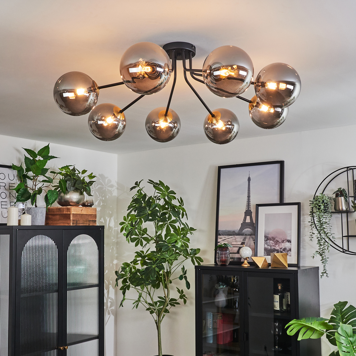 Eine moderne Deckenlampe mit kugelförmigen Schirmen und Pflanzen. Ein schwarzer Schrank und ein gerahmtes Eiffelturm-Bild.