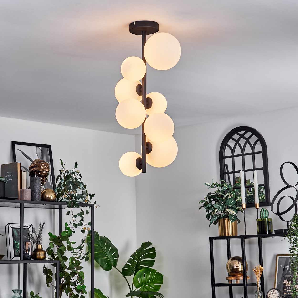 Eine moderne Deckenleuchte mit mehreren kugelförmigen Lichtern hängt in einem Raum. Regale mit Pflanzen und Dekoration sind im Hintergrund sichtbar.