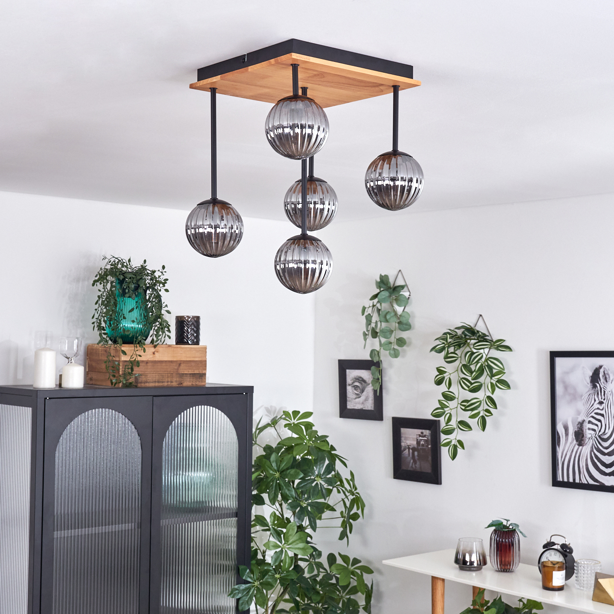 Modernes Interieur mit Deckenleuchte, Pflanzen und Möbeln. Ein Zebrastreifen und gerahmte Bilder schmücken eine weiße Wand.