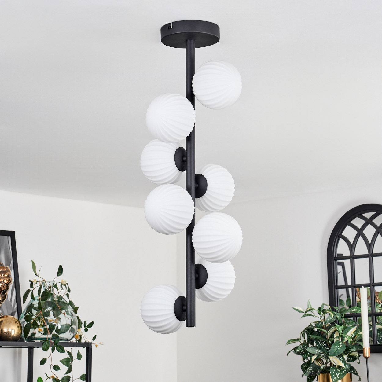 Eine moderne Deckenleuchte mit weißen Kugelleuchten hängt von der Decke. Schwarze Metallakzente. Pflanzen und Dekoration sind im Hintergrund sichtbar.