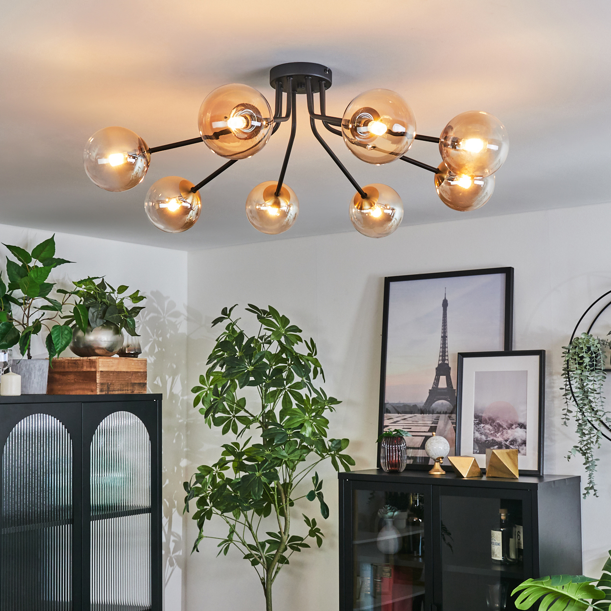 Eine moderne Deckenleuchte mit kugelförmigen Lichtern hängt über Pflanzen und Dekoration. Kunstwerke des Eiffelturms und einer rosa Landschaft sind sichtbar.