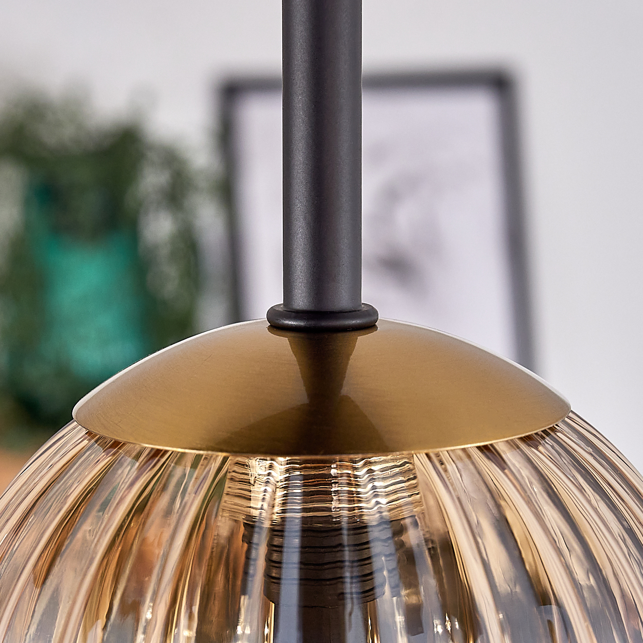 Nahaufnahme einer goldenen und gläsernen Lampe. Ein dunkler Stab trägt die Lampe. Im Hintergrund eine verschwommene Pflanze und ein gerahmtes Bild.