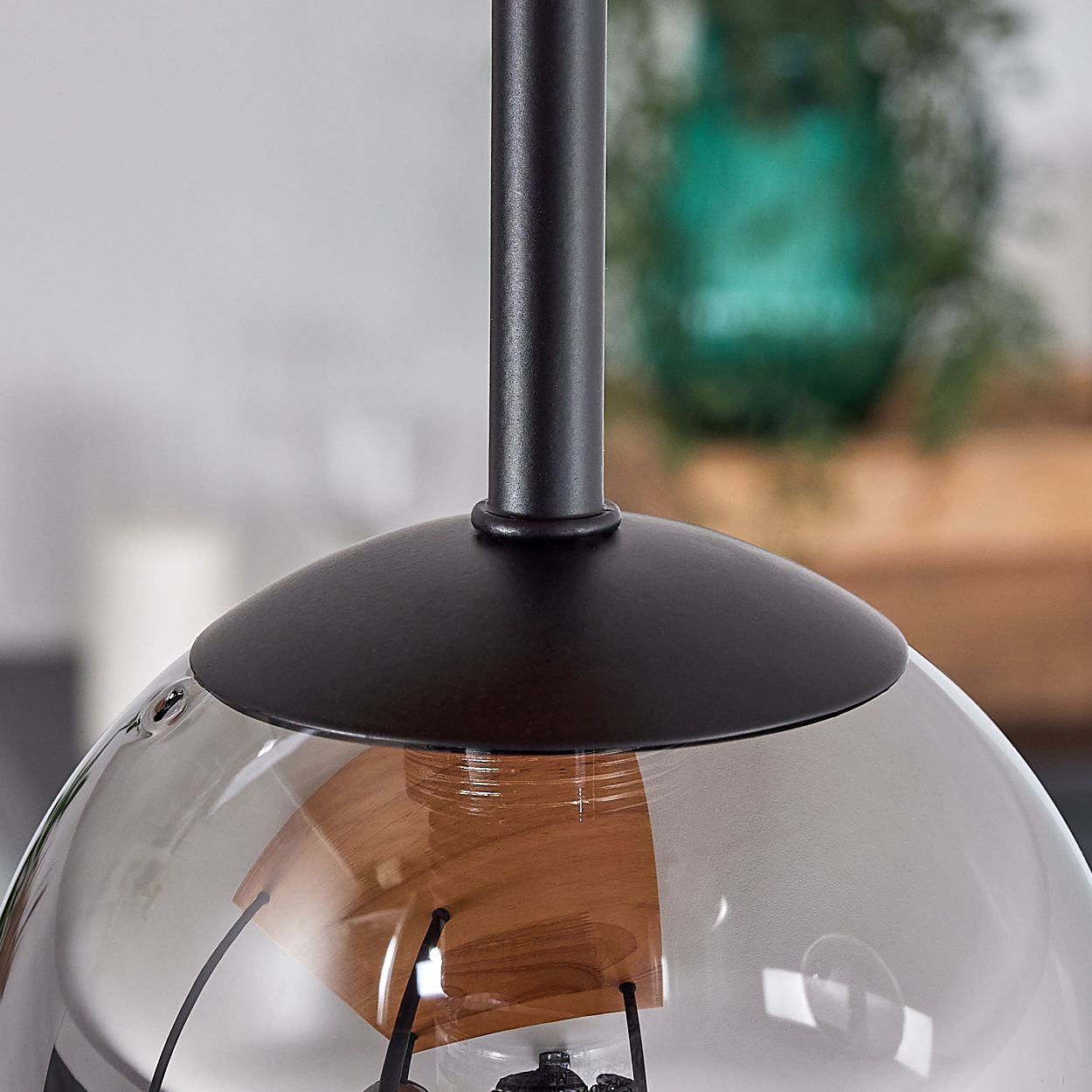 Nahaufnahme einer modernen Pendelleuchte. Sie hat einen schwarzen Metallschaft und einen klaren Glasschirm. Ein Holzelement ist sichtbar.
