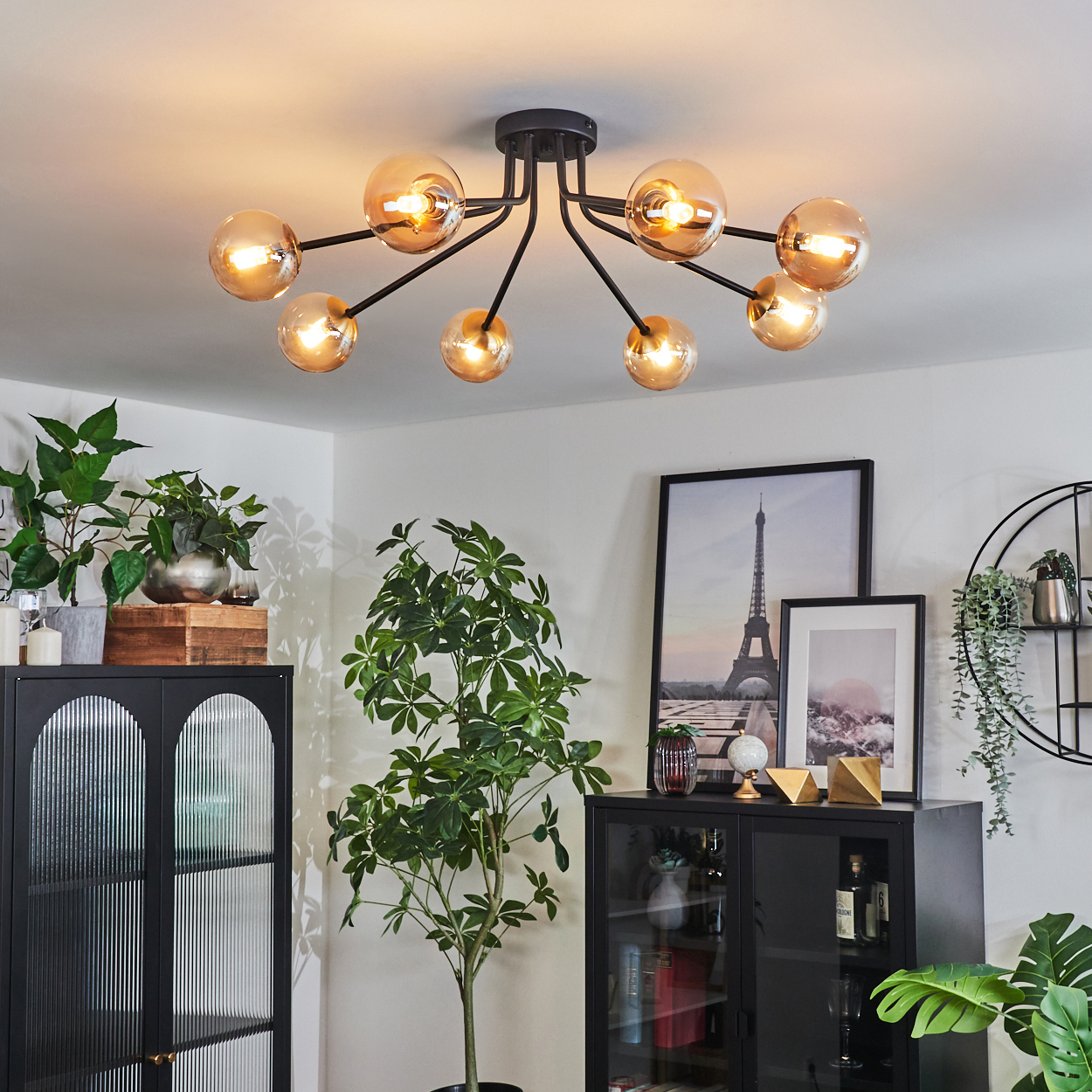 Ein modernes Interieur mit einem schwarzen Kronleuchter, Pflanzen und gerahmten Bildern. Schwarze Schränke stehen im Vordergrund, die Decke ist weiß.