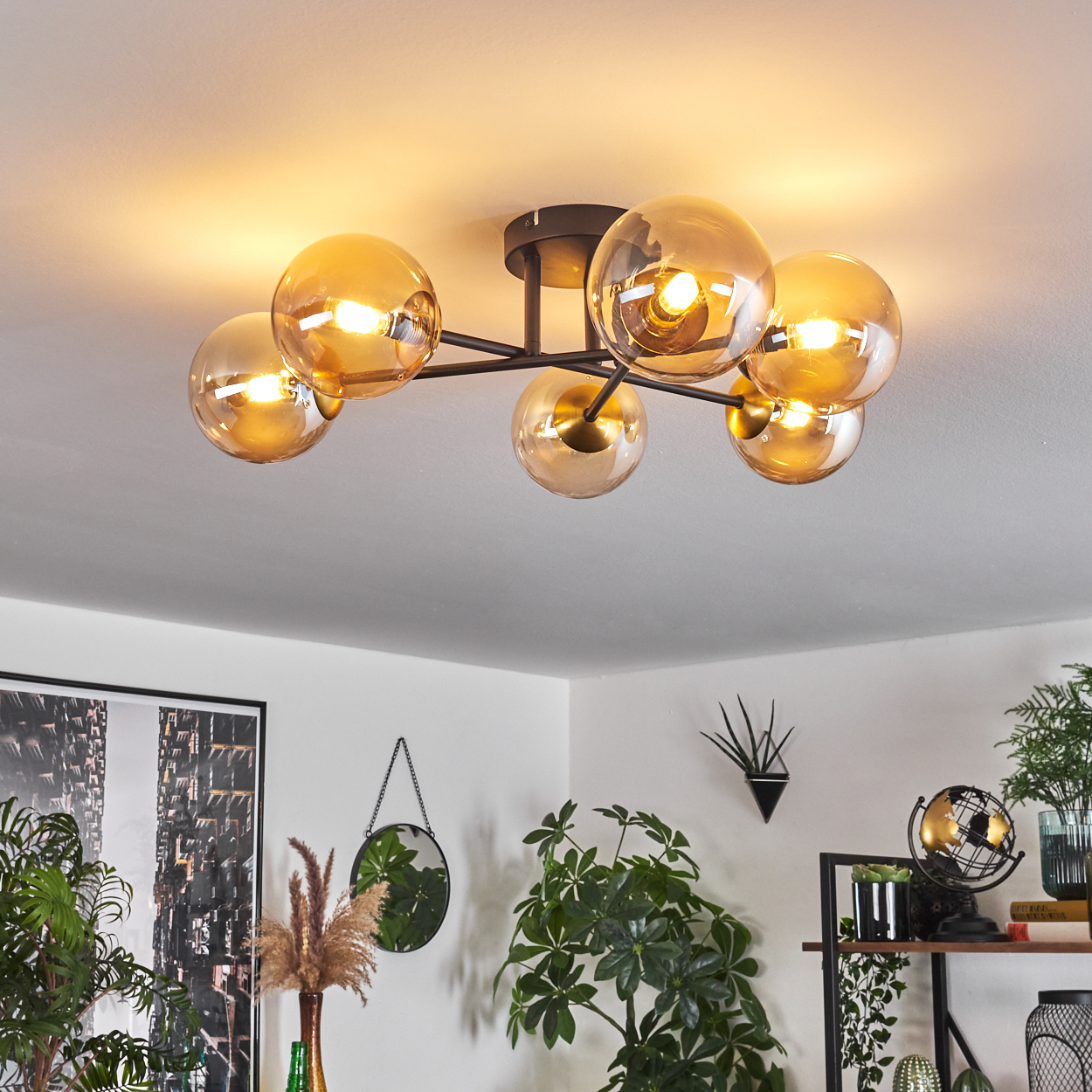 Eine Deckenlampe mit sechs bernsteinfarbenen Glaskugeln beleuchtet einen Raum. Im Hintergrund sind Pflanzen und Dekorationen zu sehen.