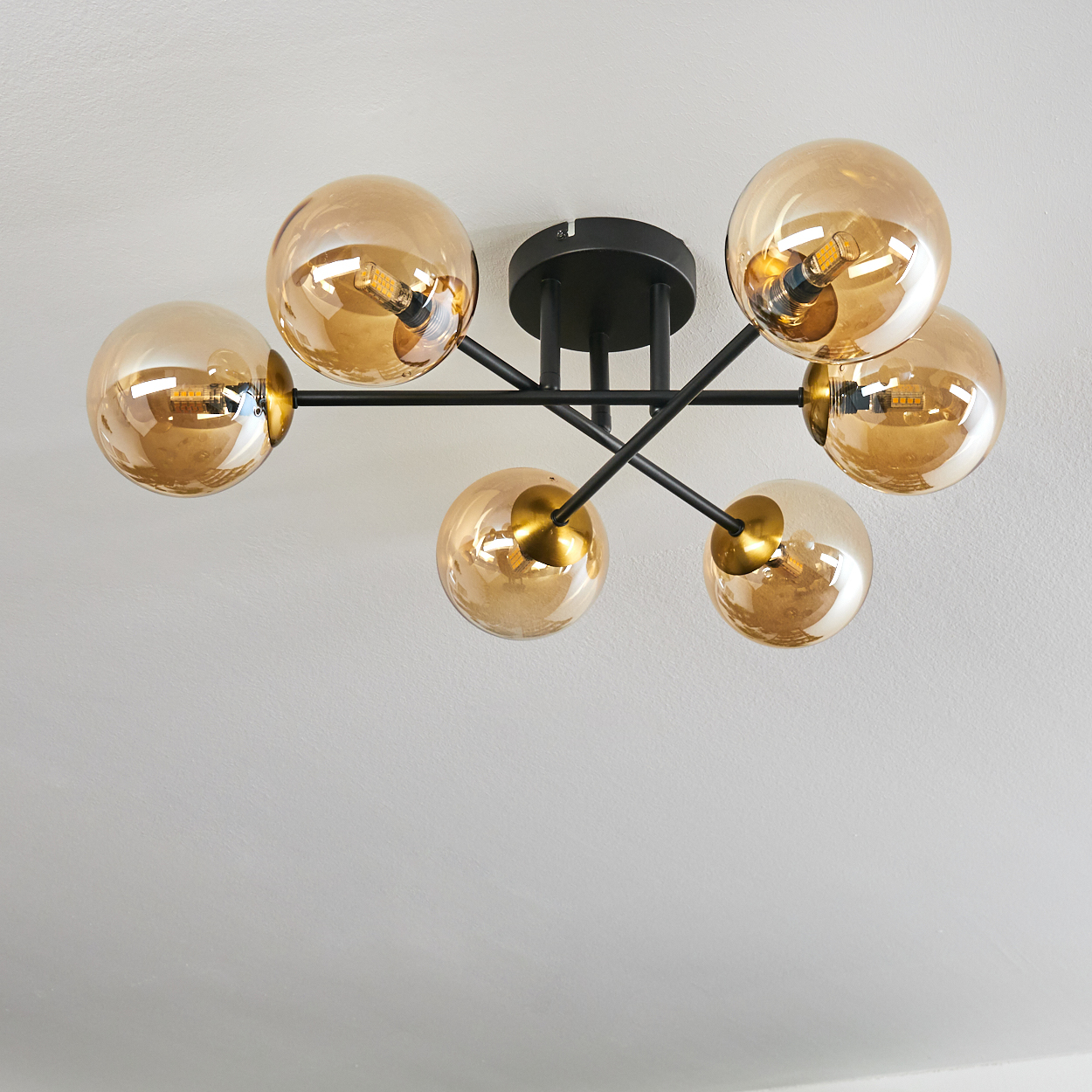 Eine moderne Deckenleuchte mit sechs bernsteinfarbenen Glaskugeln. Sie ist an einer weißen Decke montiert. Die Leuchte hat schwarze Metallarme.