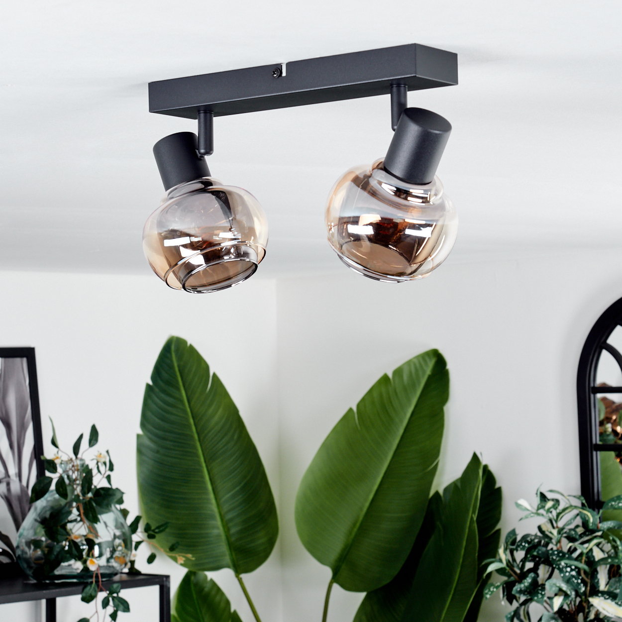 Zwei-Licht-Leuchte mit bernsteinfarbenen Glaskugeln, schwarzem Rahmen, über großen grünen Blättern und einer Vase mit Blumen.