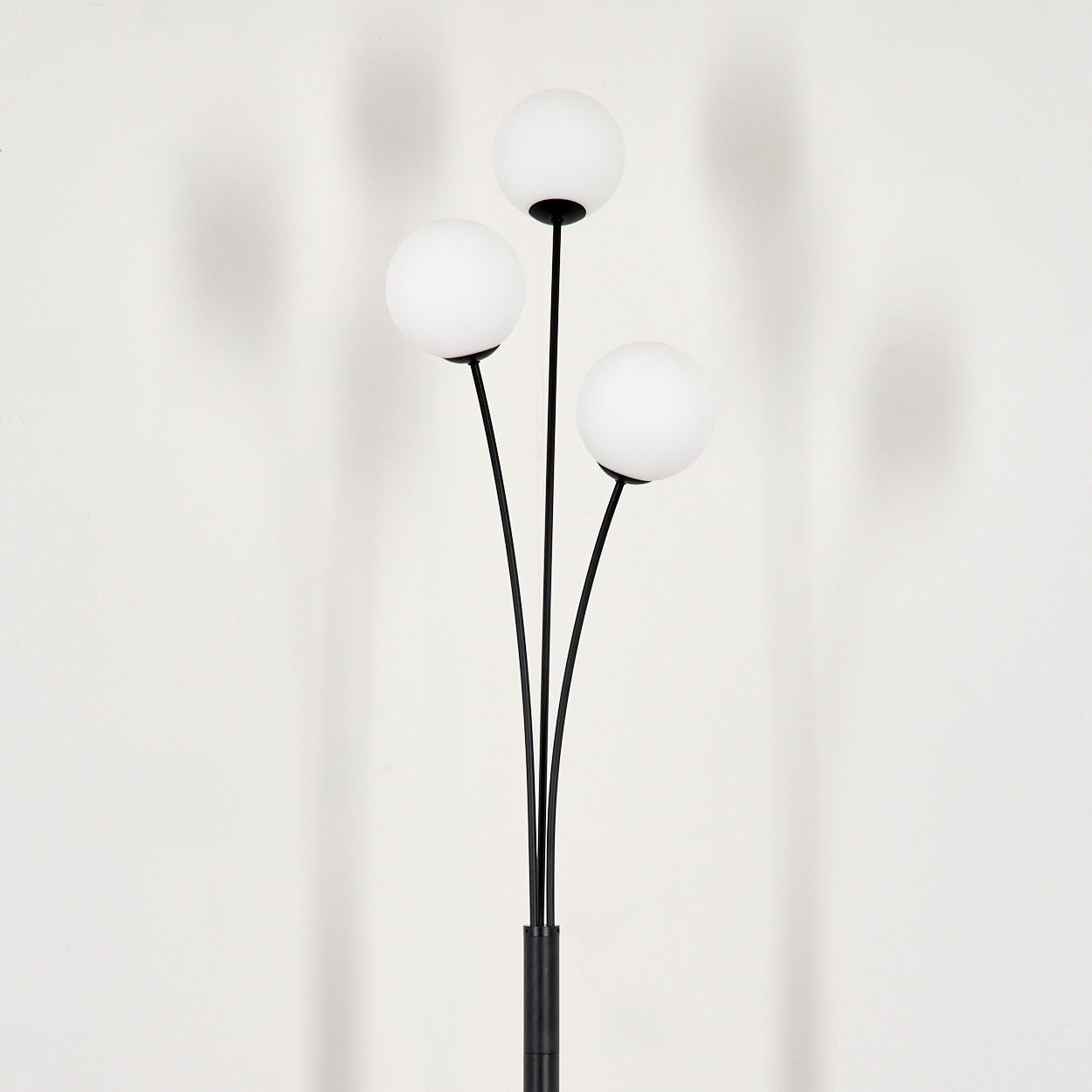 Eine schwarze Stehlampe mit drei weißen Kugelleuchten. Die Lampe steht vor einer weißen Wand, und dahinter sind Schatten zu sehen.