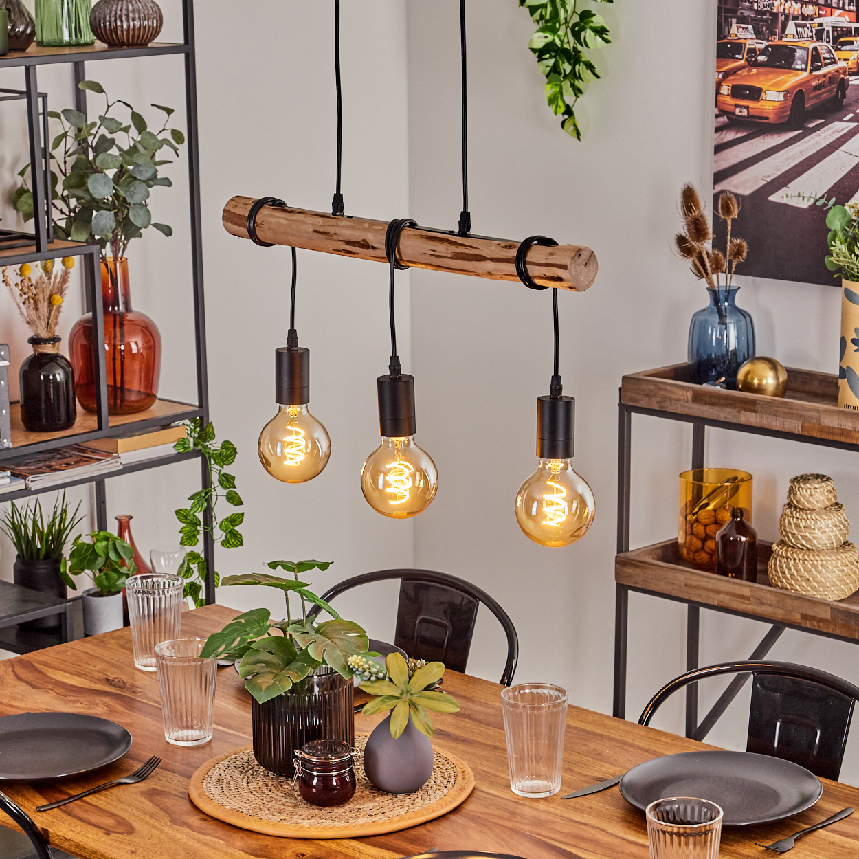 Ein Esszimmer mit Holztisch, schwarzen Stühlen und einer Hängelampe mit drei Glühbirnen. Dekorative Gegenstände auf Regalen.