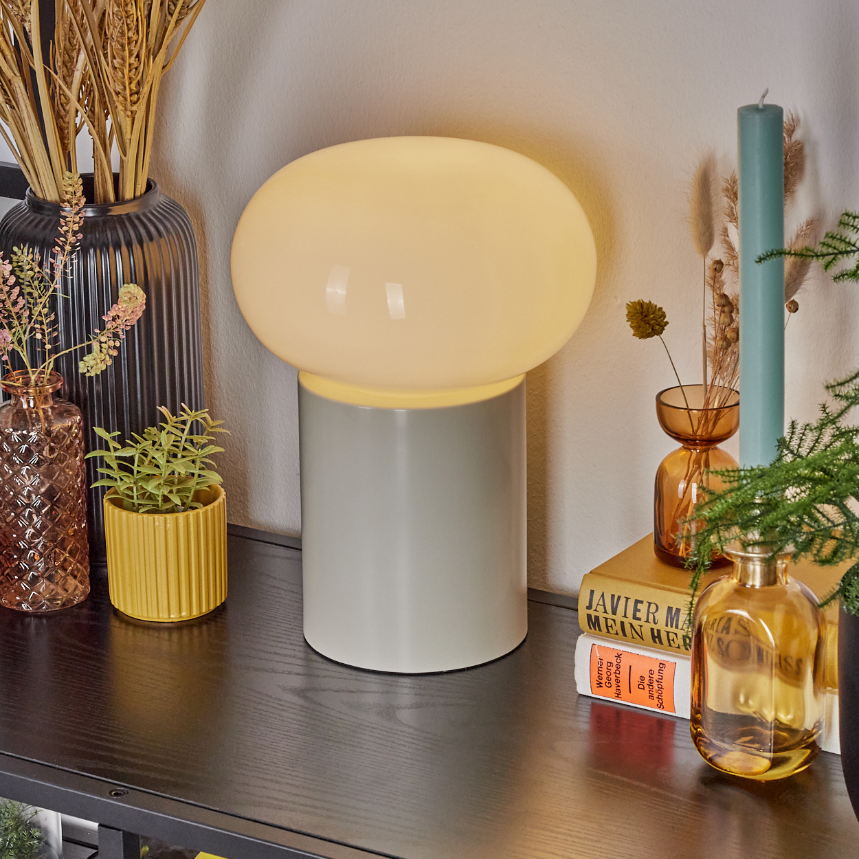 Eine beleuchtete Pilzlampe auf grauem Sockel steht auf einem schwarzen Tisch. Vasen mit Pflanzen und eine Kerze sind darum herum.