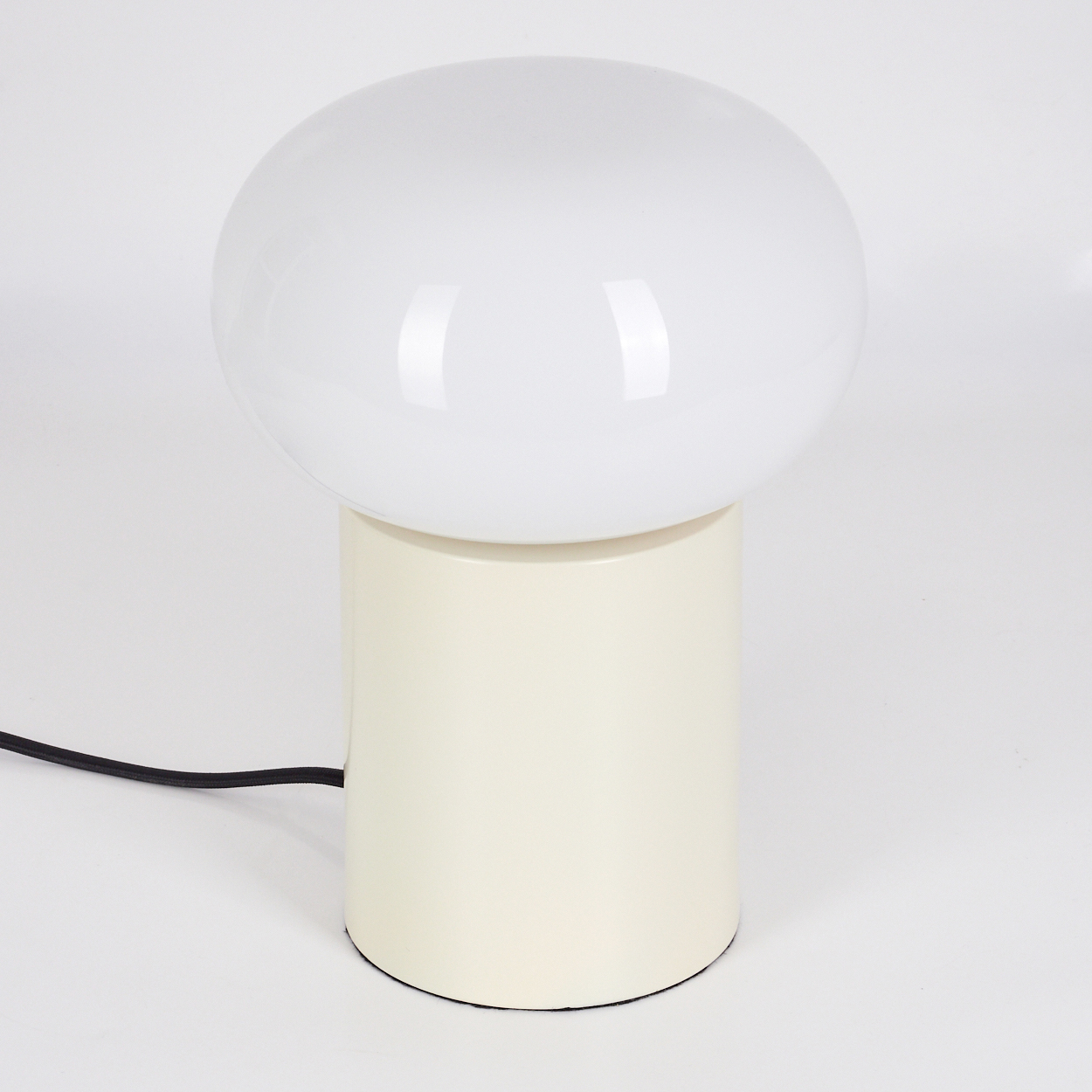 Ein weißer, kugelförmiger Lampenschirm sitzt auf einem cremefarbenen, zylindrischen Sockel. Ein schwarzes Netzkabel geht von der Lampe aus.