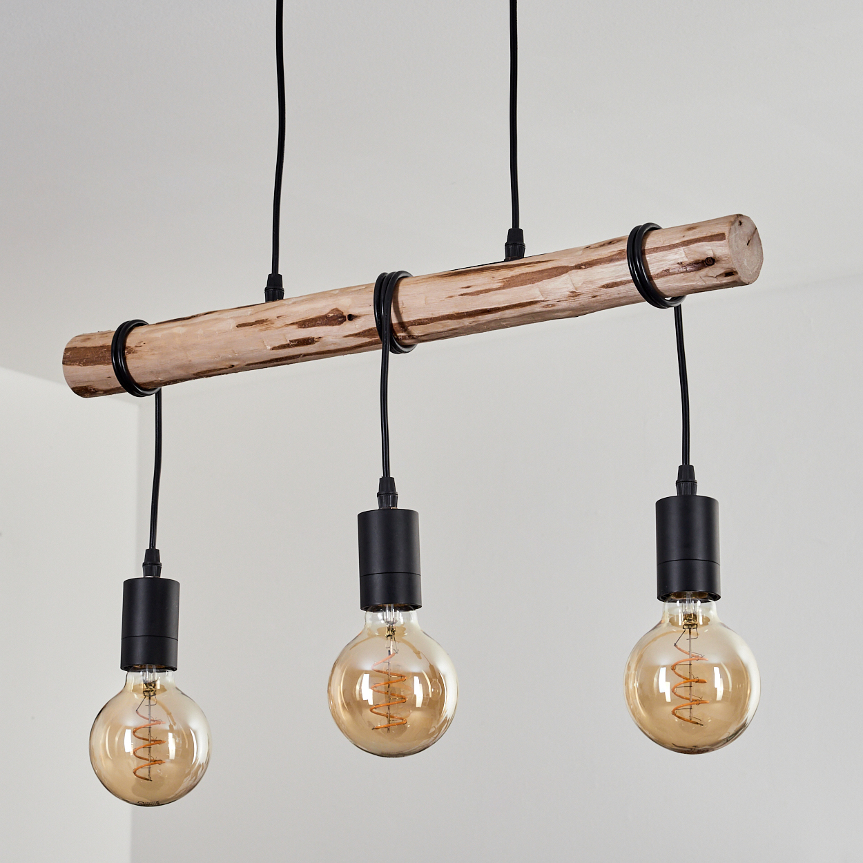 Holzbalken mit drei Pendelleuchten. Schwarze Kabel und Lampenfassungen. Warme Glühbirnen mit sichtbaren Filamenten. Weißer Wandhintergrund.