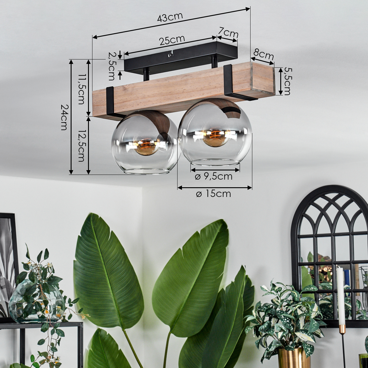Eine Deckenlampe mit zwei Glaskugeln ist an einem Holzbalken befestigt. Grüne Pflanzen und ein schwarzer Bogenförmiger Spiegel sind im Hintergrund.