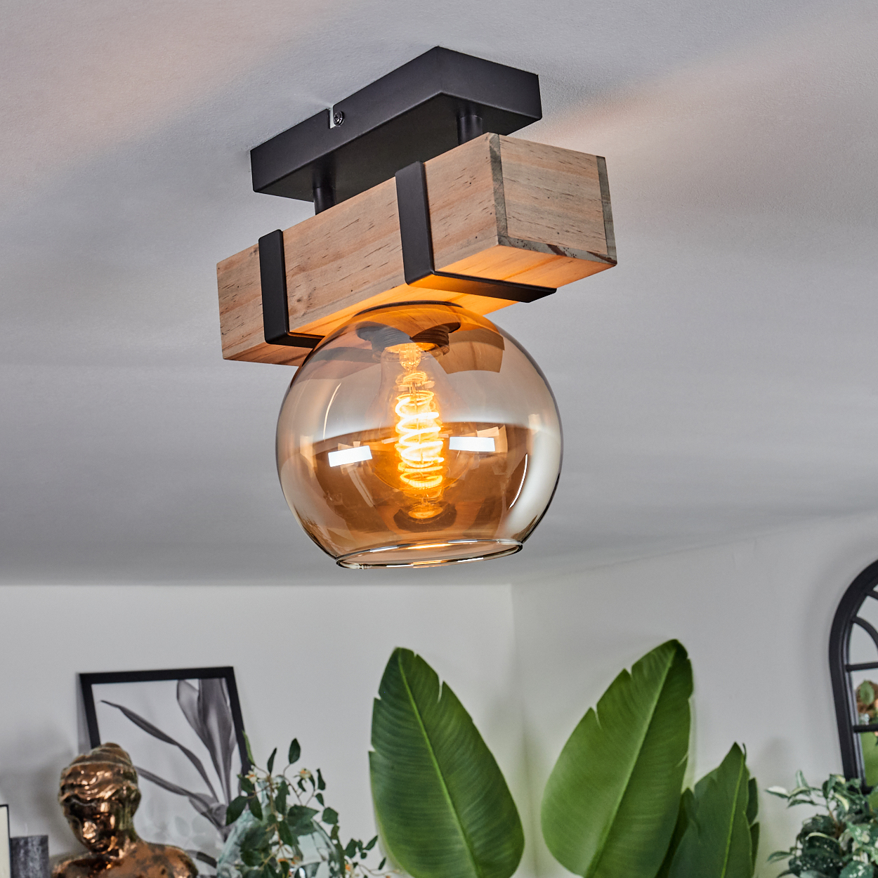 Eine Deckenlampe mit Holzbalken, schwarzem Metall und einer Glaskugel. Die Glühbirne leuchtet. Grüne Blätter und Kunst sind sichtbar.