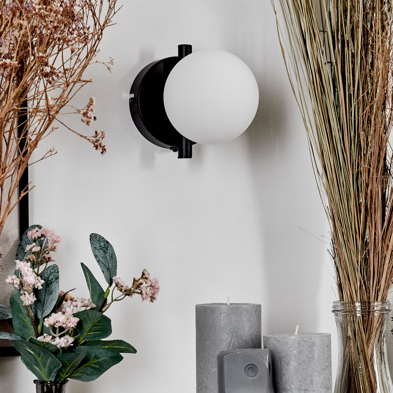 Eine moderne Wandlampe mit weißem Kugelschirm ist an einer weißen Wand montiert. Getrocknete Pflanzen, Kerzen und Blumen sind davor angeordnet.