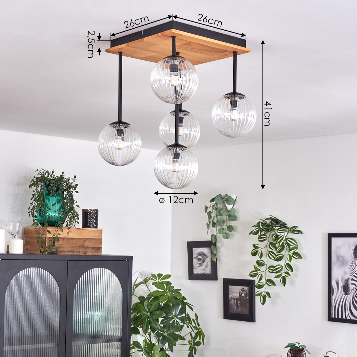 Eine Deckenlampe aus Holz mit fünf Glaskugeln hängt in einem Raum mit Pflanzen und gerahmten Bildern. Abmessungen sind angegeben.