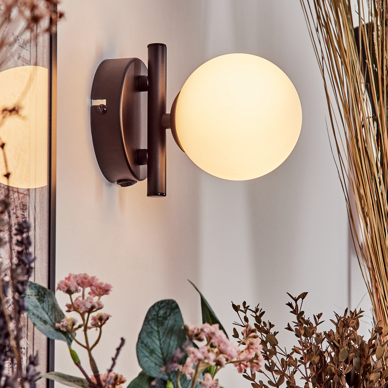 Eine moderne Wandleuchte mit rundem, leuchtendem Licht. Braune Metallhalterung. Blumen und getrocknete Pflanzen umrahmen das Licht.