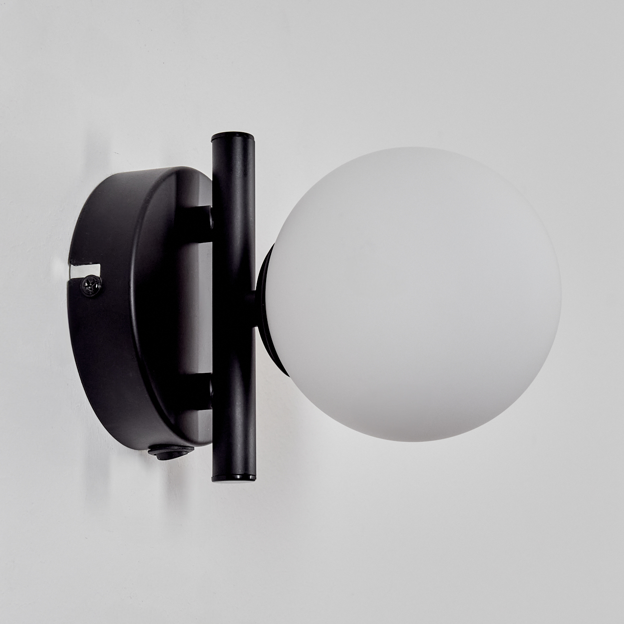 Eine schwarze Wandlampe mit kugelförmigem, weißem Lampenschirm ist an einer weißen Wand montiert. Der Arm der Lampe ist vertikal.