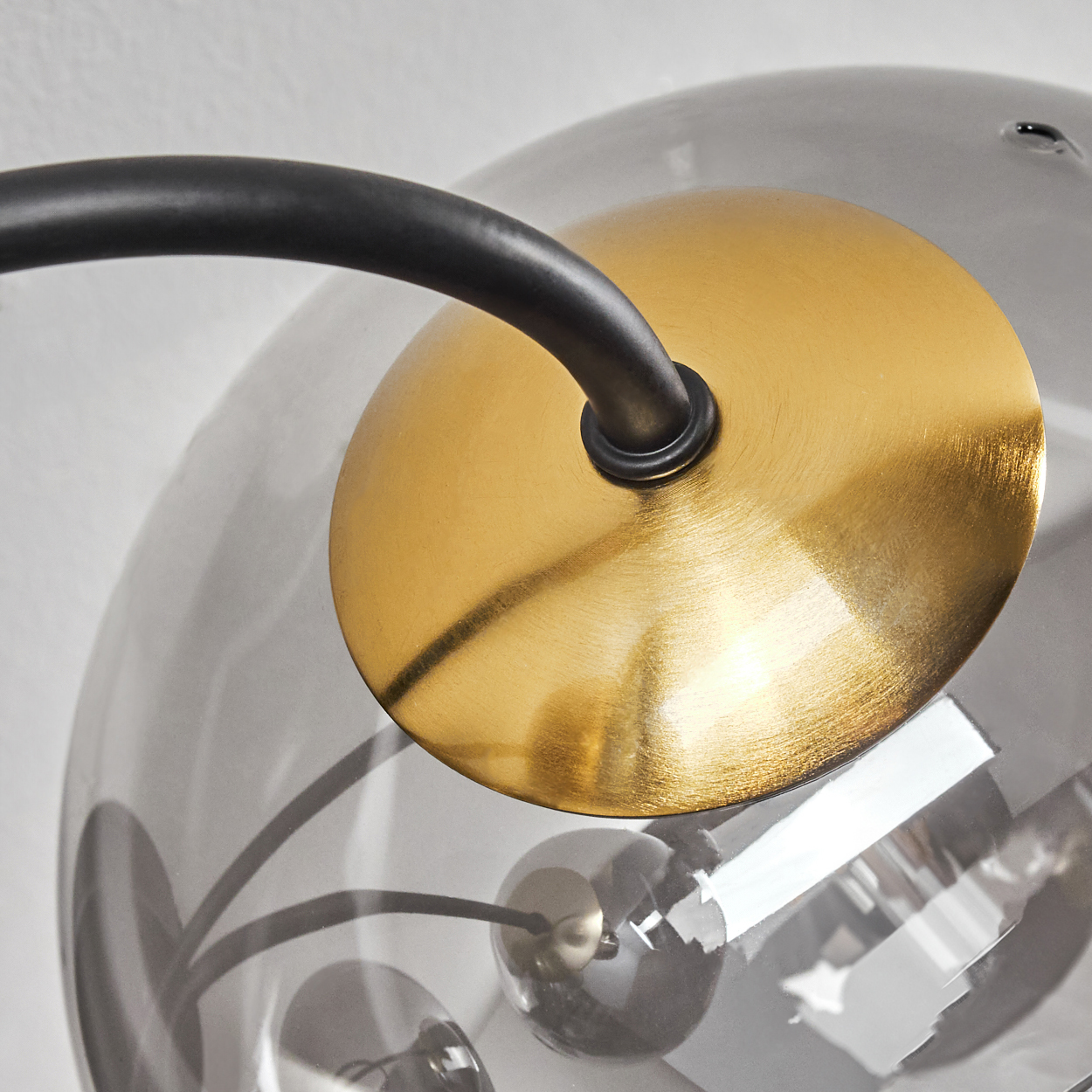 Nahaufnahme einer Lampe. Sie hat einen schwarzen, gebogenen Arm, eine goldene Scheibe und eine rauchige Glaskugel. Der Hintergrund ist eine weiße Wand.