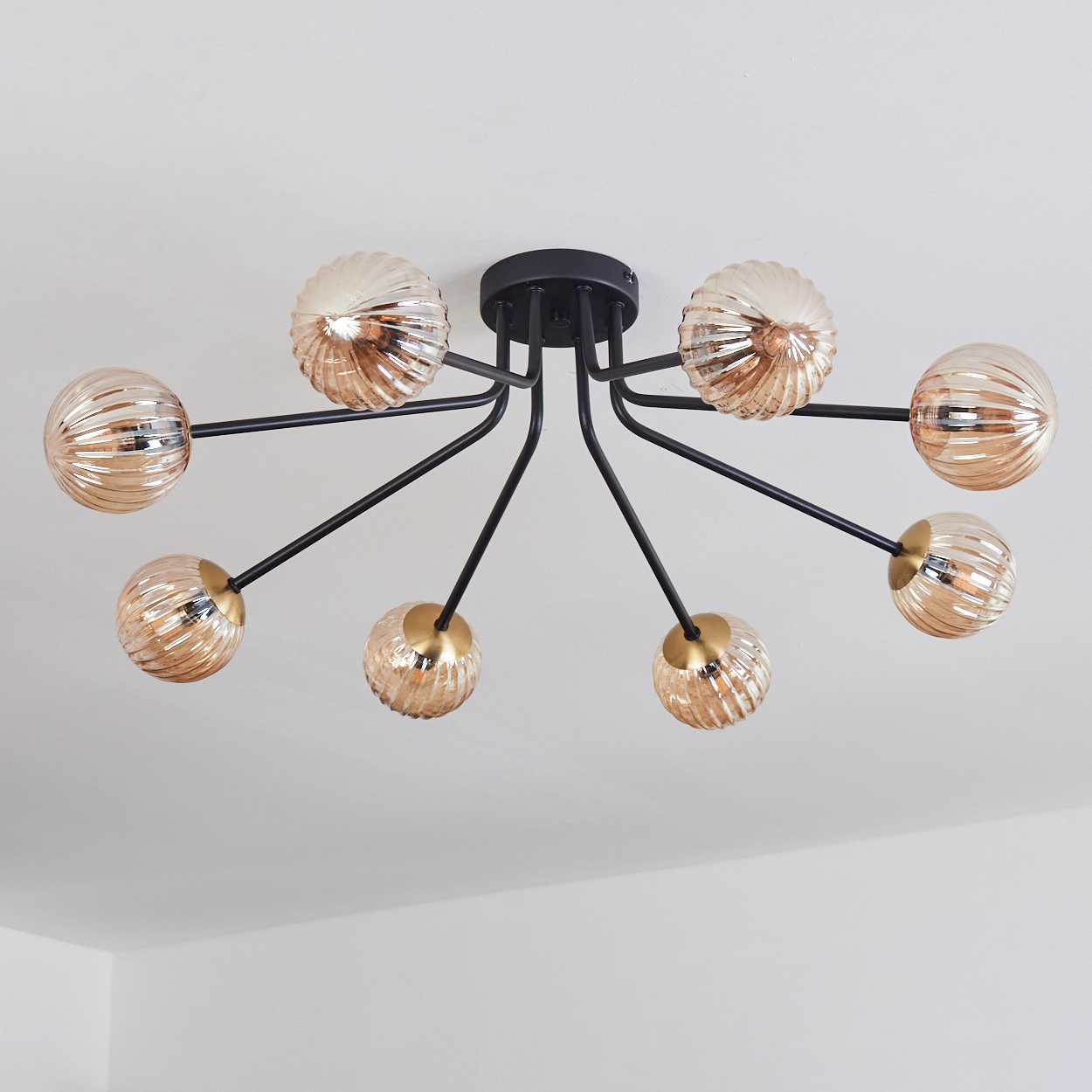 Eine moderne Deckenleuchte mit acht bernsteinfarbenen Glaskugeln und schwarzen Metallarmen, die an einer weißen Decke montiert ist.